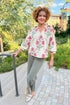 Woman in a floral blouse and gray pants standing on a sidewalk with greenery in the background