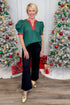 Front view of woman wearing green top and black jeans with Christmas trees in the background.