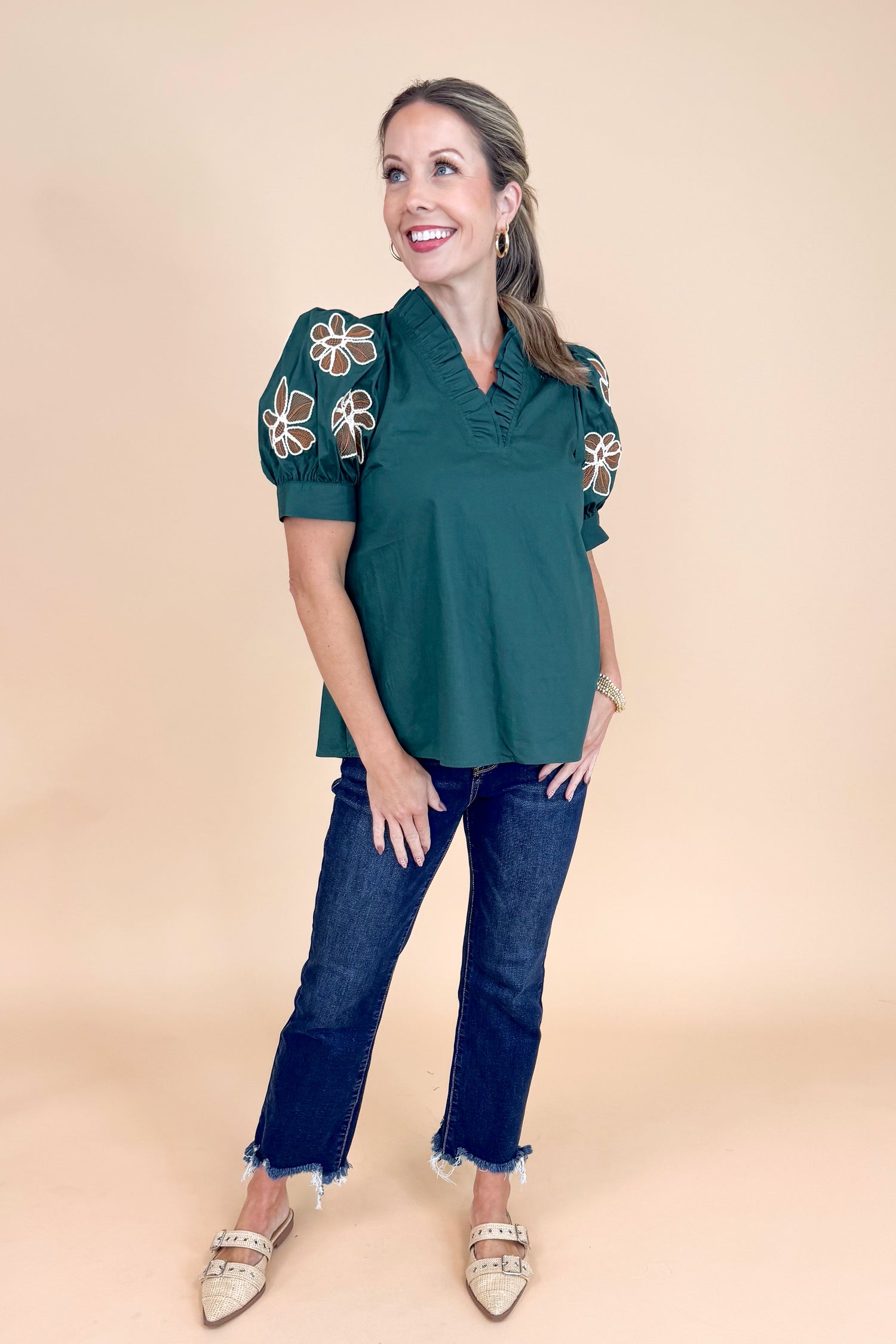 Woman wearing a dark green top with floral embroidery and jeans on a beige background