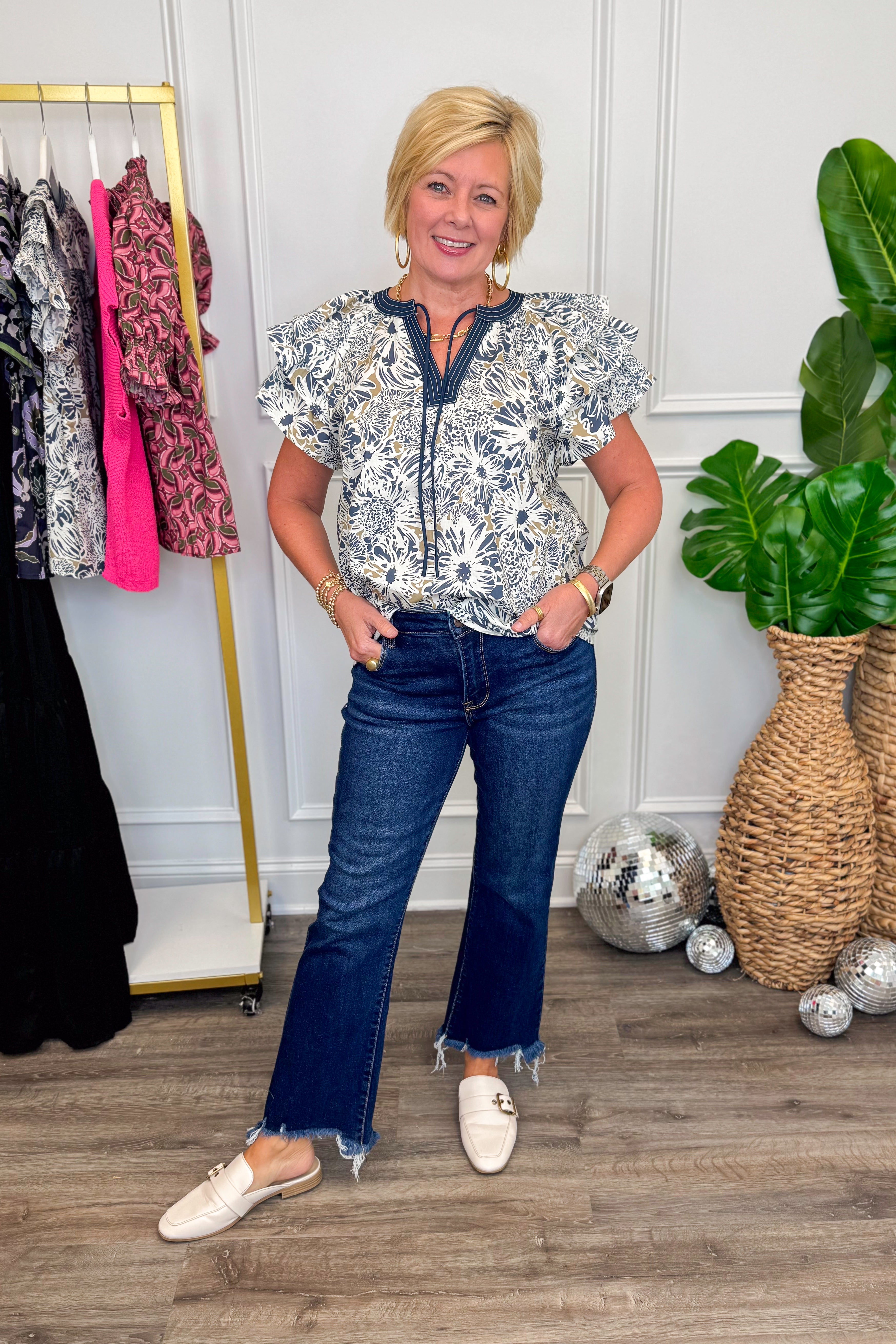 Woman in a patterned blouse and blue jeans standing in a room with a rack of clothes and decorative items.