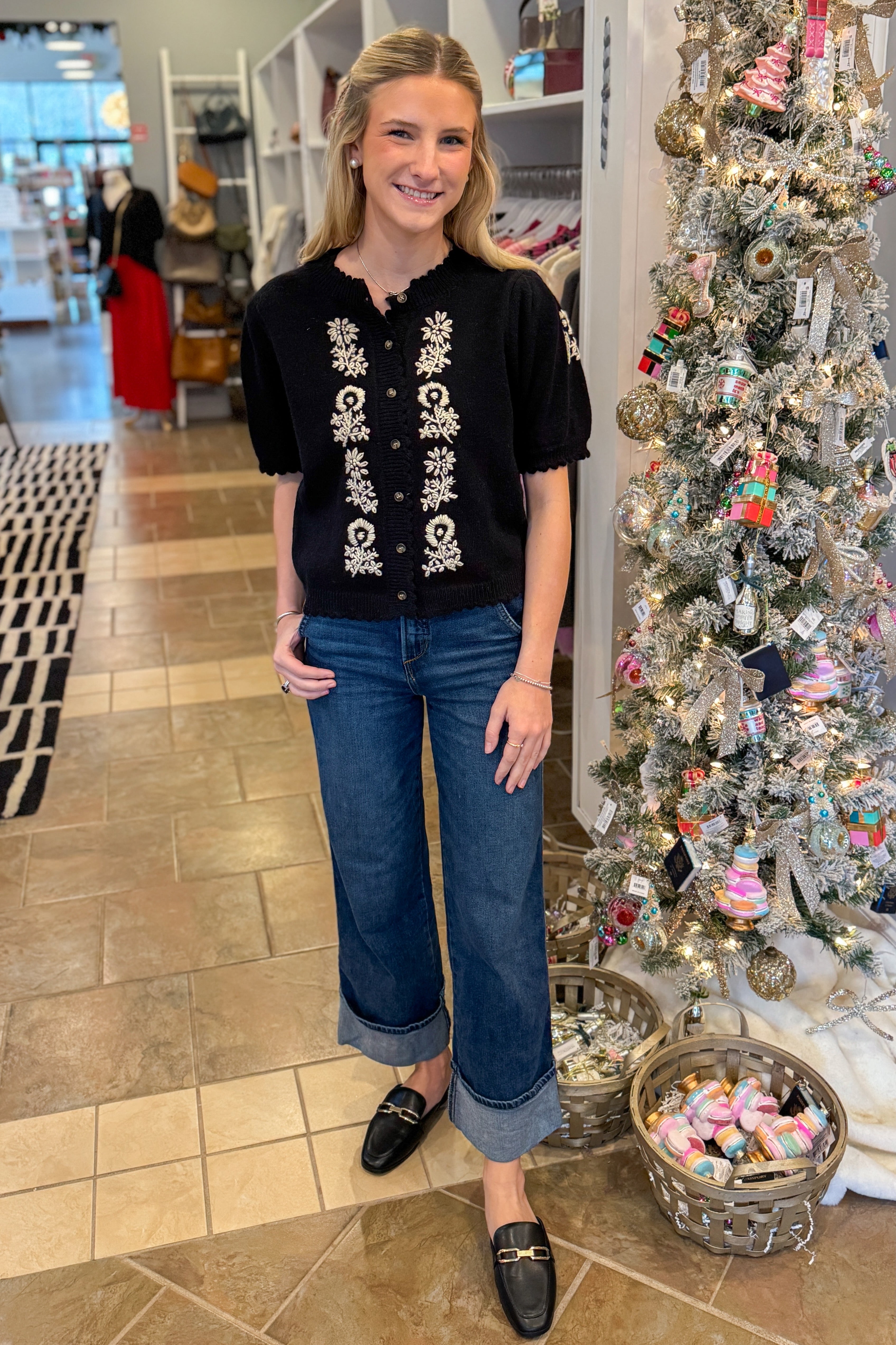 Front view of woman wearing Black Center Embroidered Short Sleeve Cardigan with jeans against holiday decor.