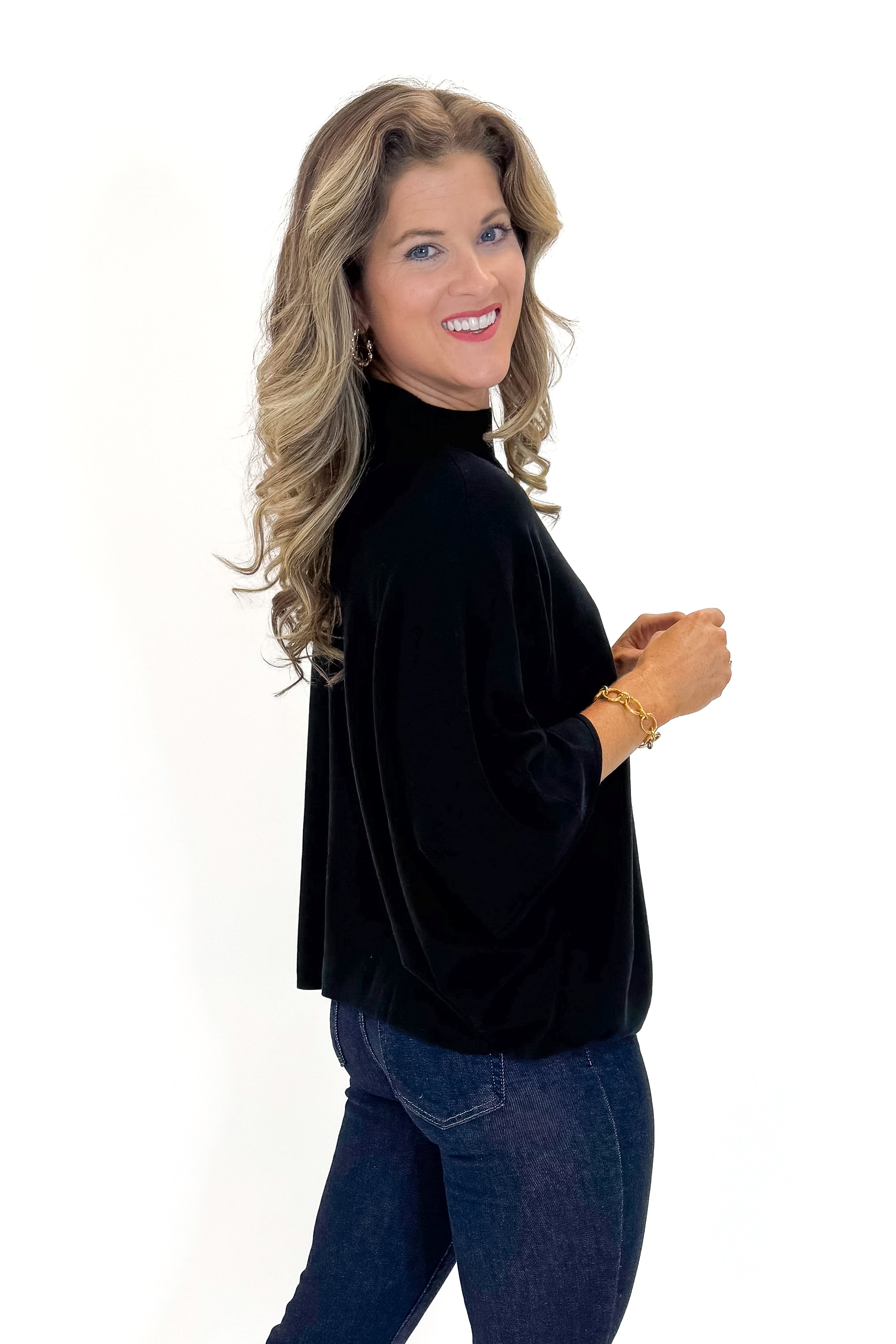 Side view of woman wearing Black Mock Neck Poncho Sweater with jeans against white studio background.
