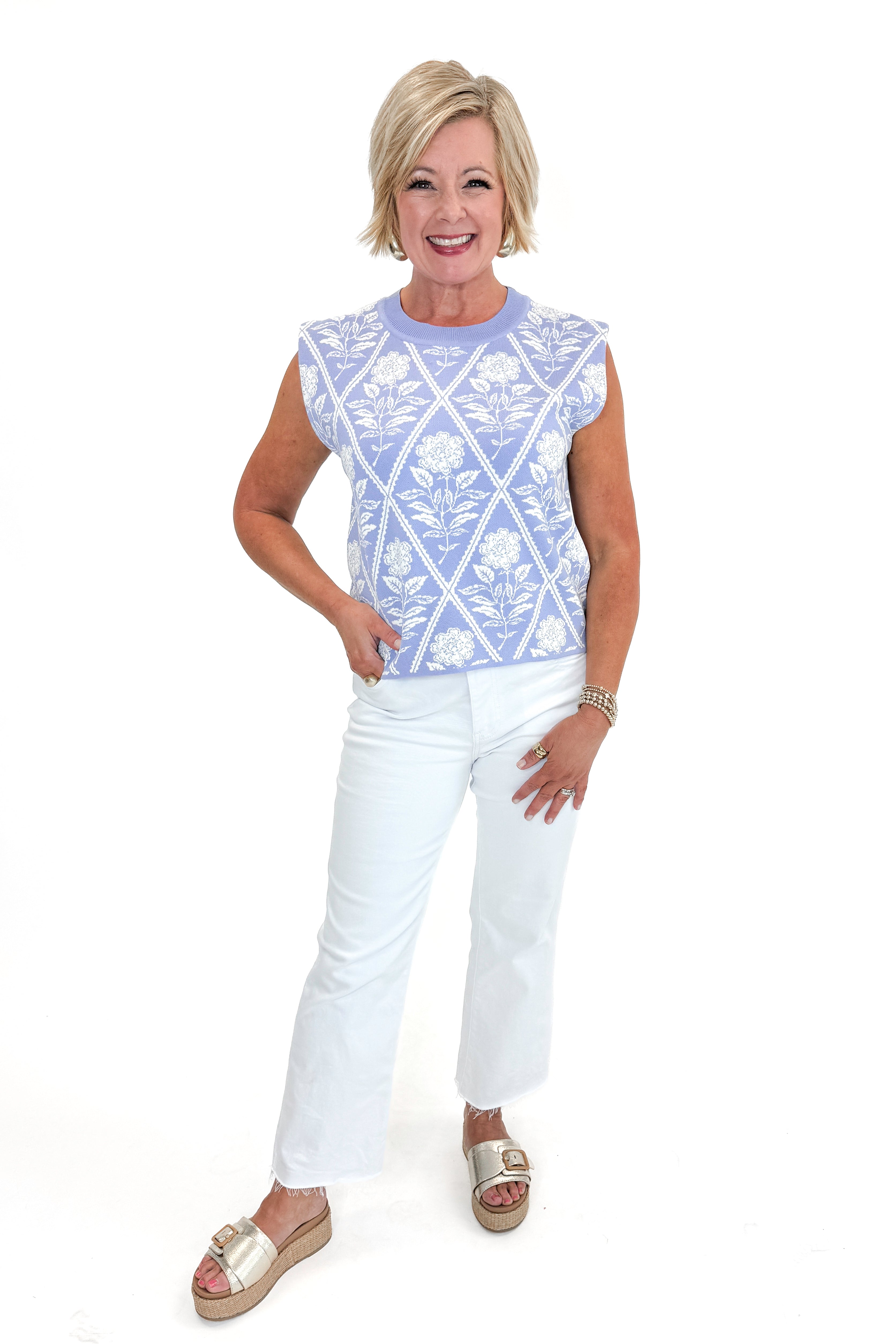 Front view of woman wearing Periwinkle Floral Printed Sleeveless Sweater by Entro with white jeans against white studio background.