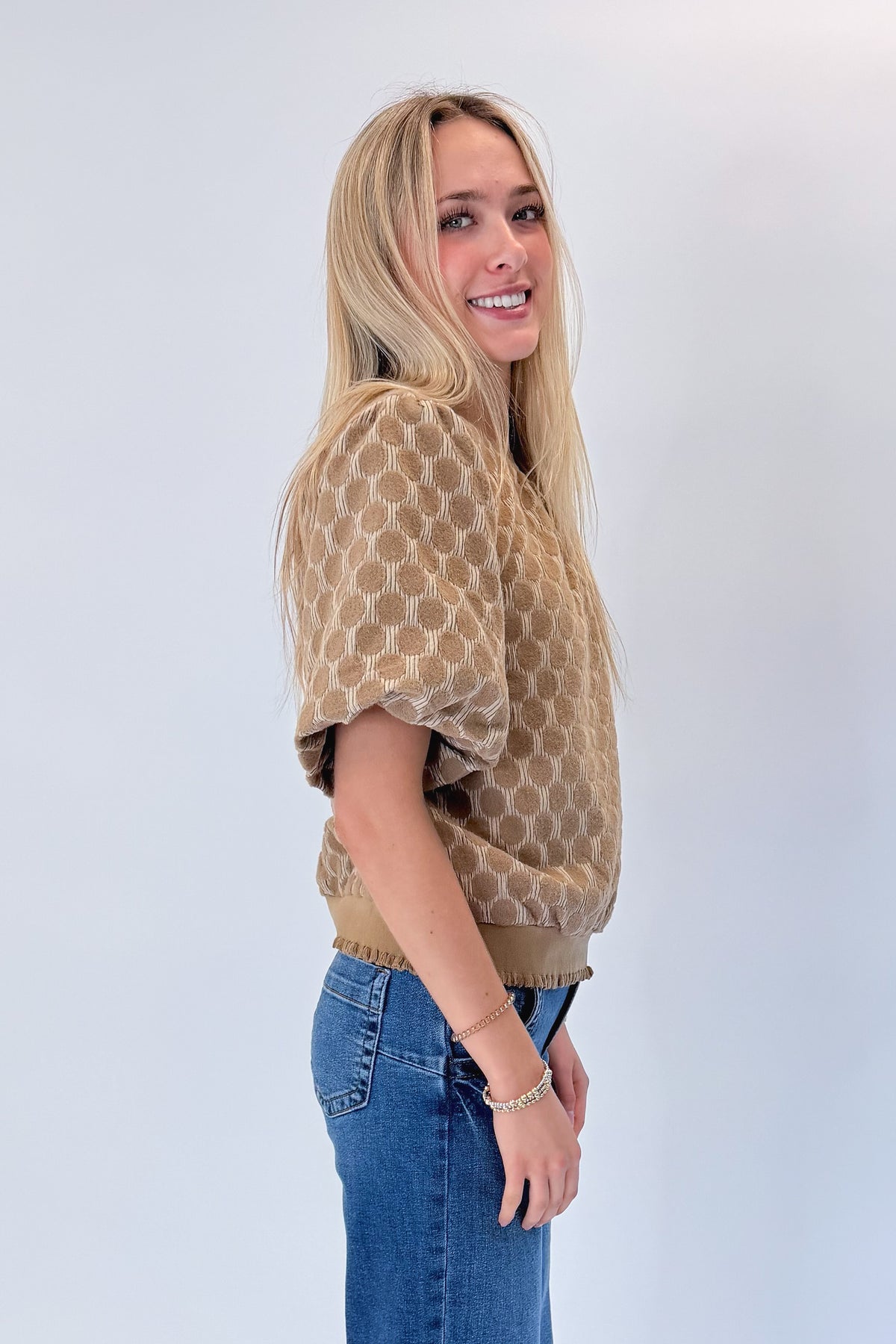 Side view of woman wearing Swagger Boutique’s Taupe Polka Dot Textured Top with jeans against white studio background.