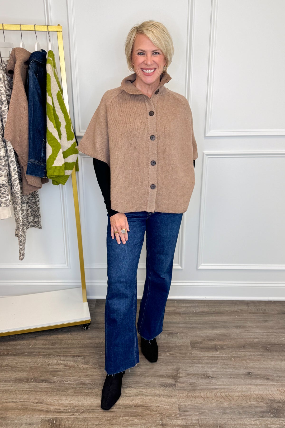 Front view of woman wearing Swagger Boutique’s Taupe Button Down Cape Jacket with jeans with clothing hanging in the background.