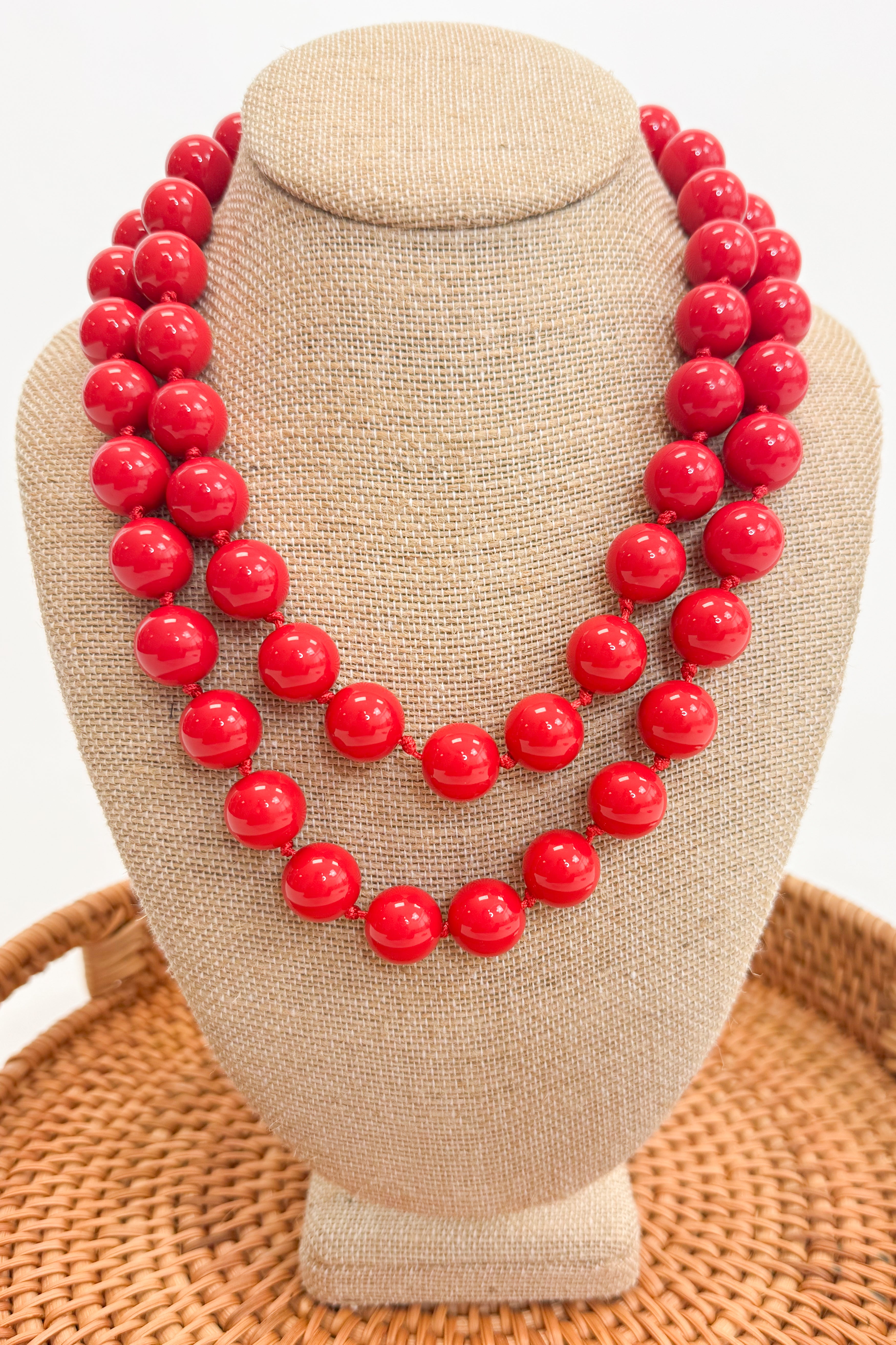 Red beaded necklace on a beige jewelry display stand with a woven surface below.
