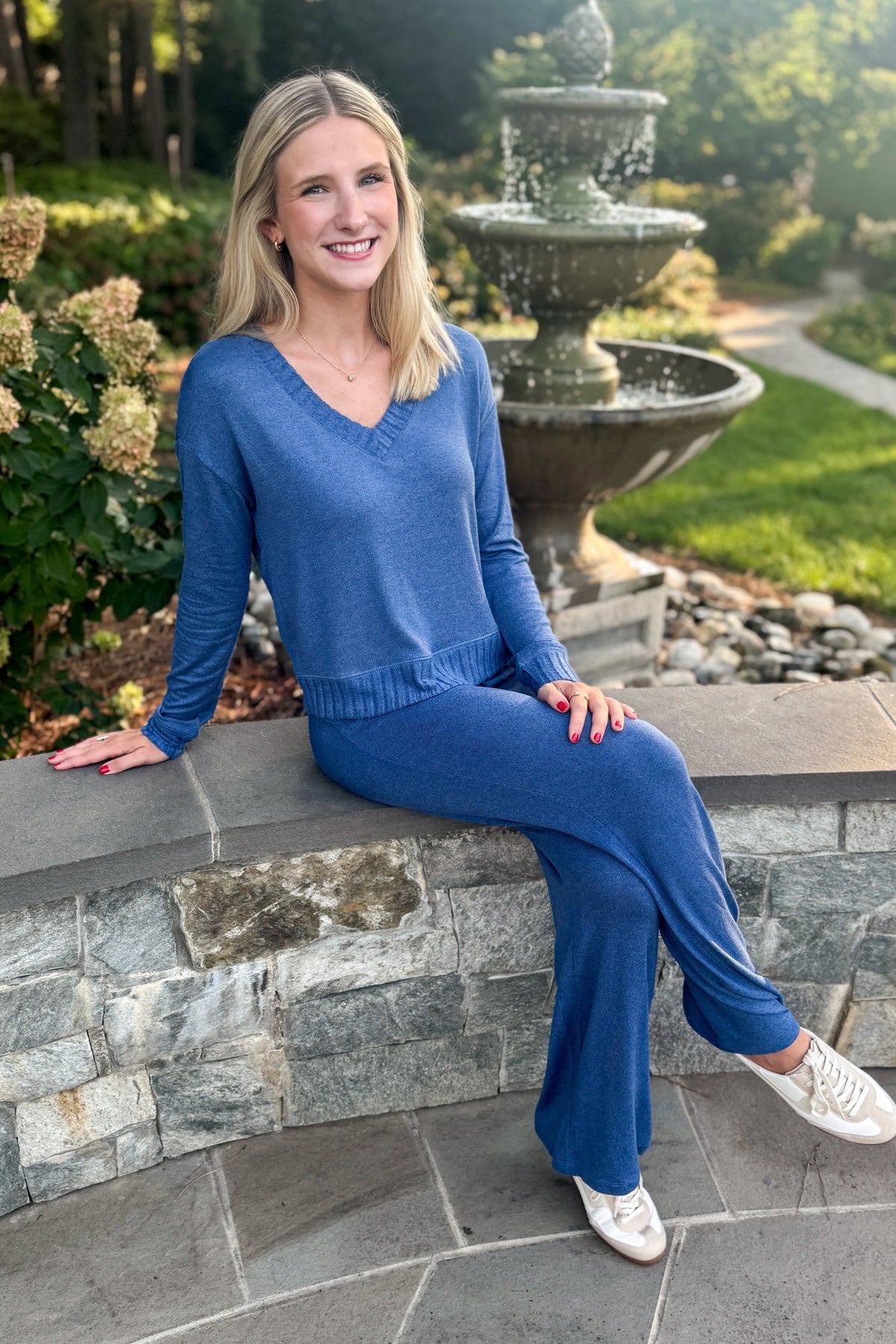 A woman wearing a matching blue knit sweater and wide-leg pants set sits on a stone wall in a garden setting with a fountain in the background.