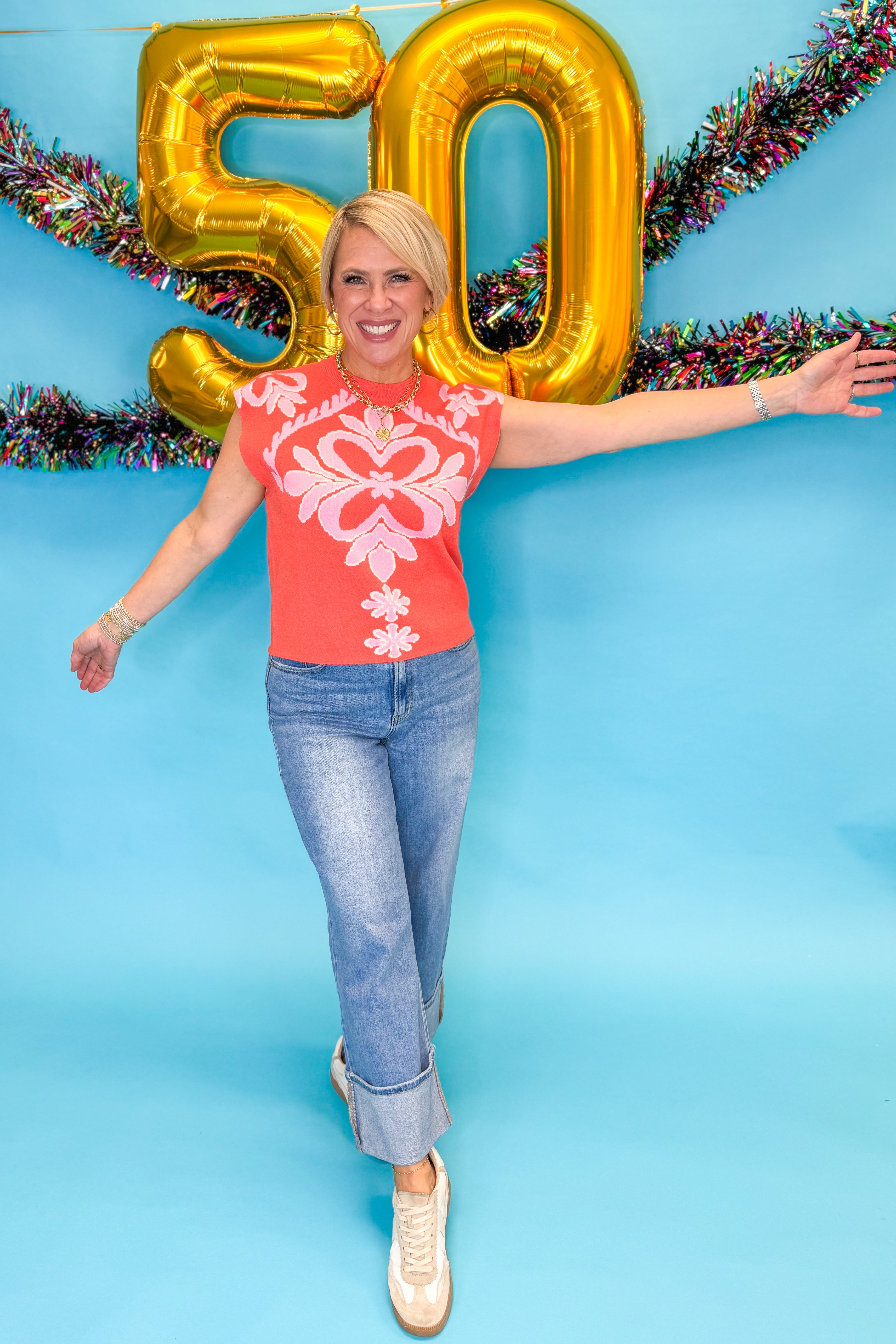Front view of woman wearing Orange & Pink Printed Sleeveless Sweater with jeans against blue background.