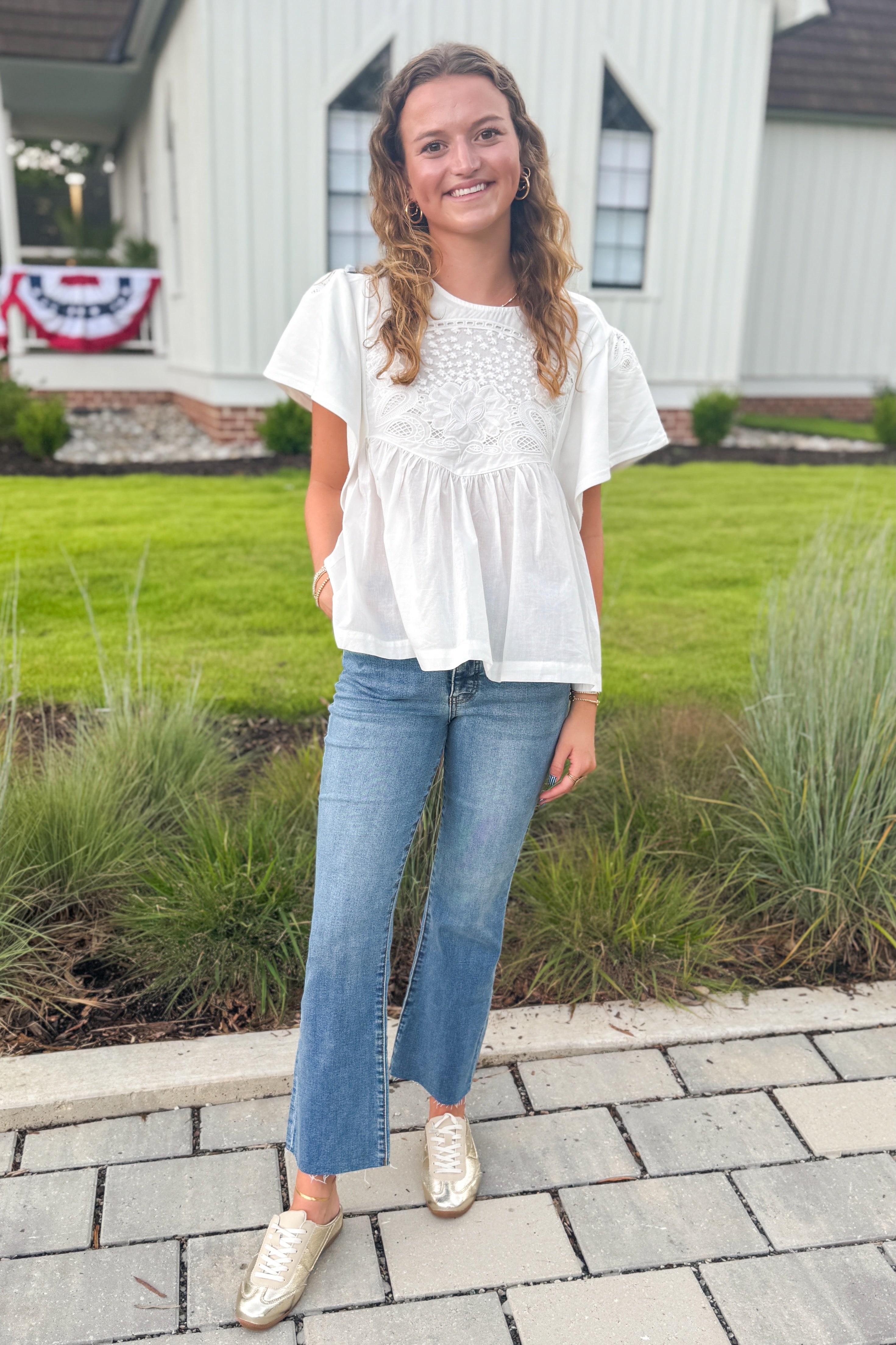 Front view of woman wearing Swagger Boutique’s White Embroidered Peplum Top with jeans standing outside by a house.