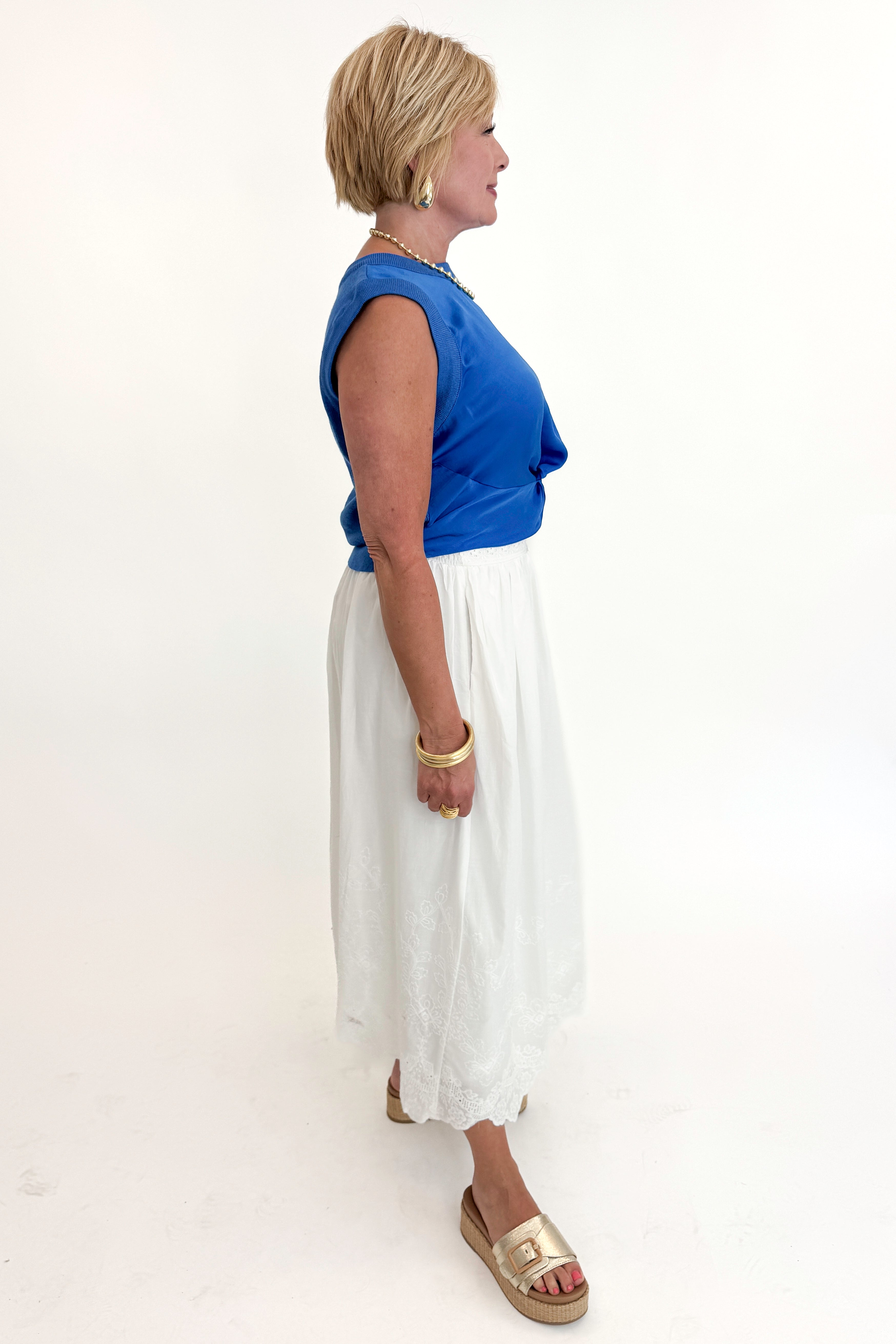 Side view of woman wearing Swagger Boutiqueβs Blue Sleeveless Tank With Twist by Skies Are Blue with White Embroidered Skirt against white studio background.
