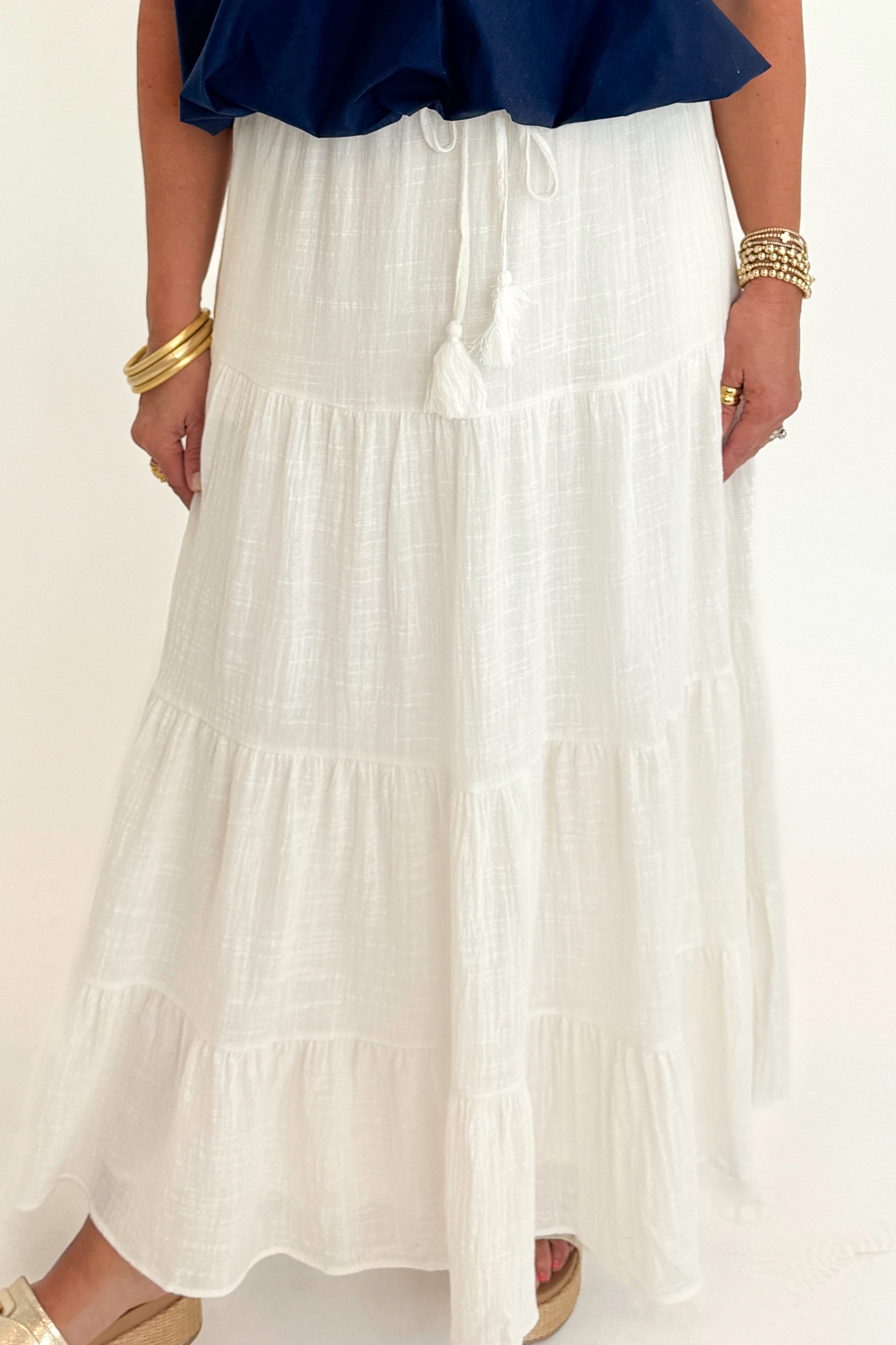 Front detail view of woman wearing Swagger Boutique’s White Flowy Tiered Cotton Skirt with blue top against white studio background.