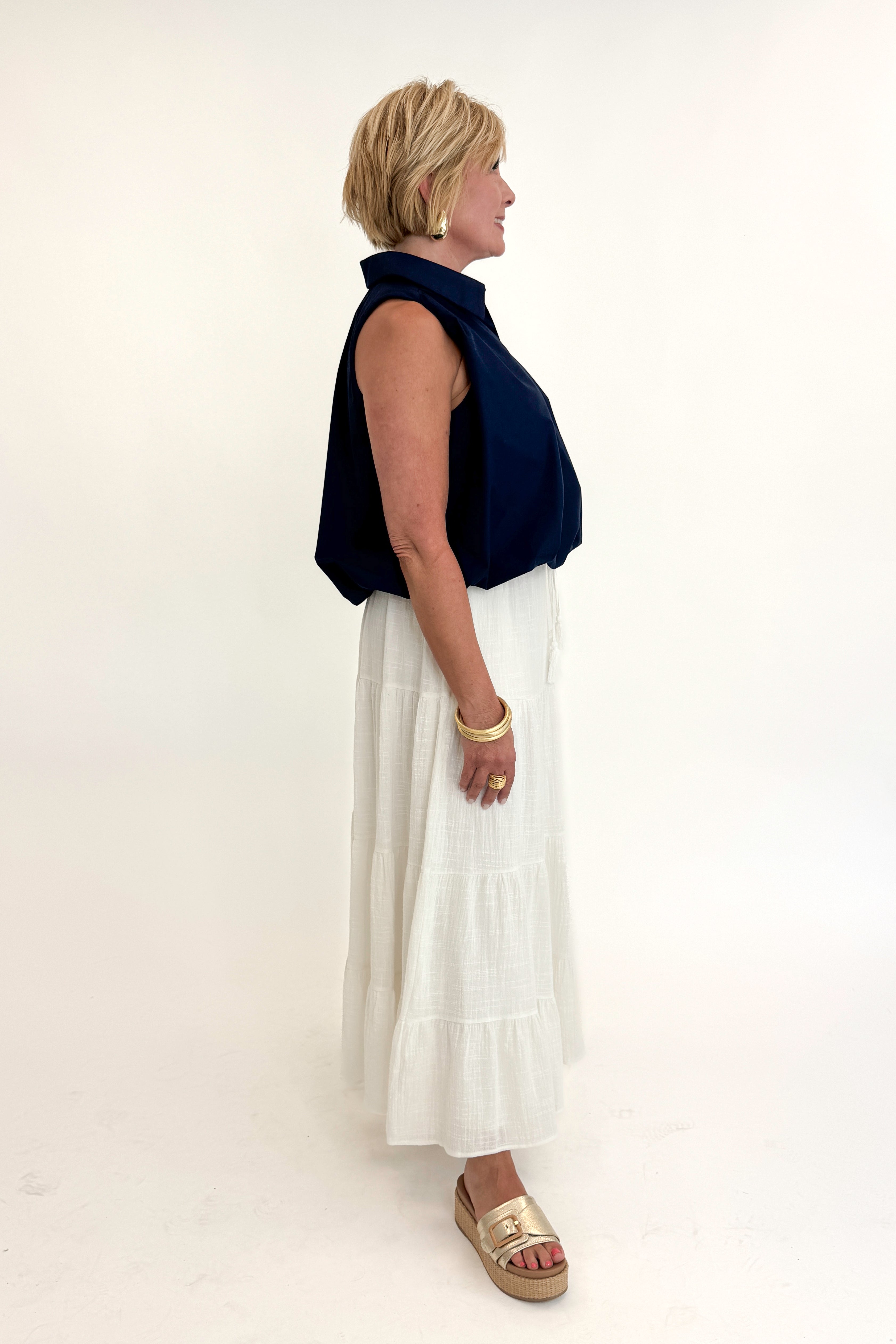 Side view of woman wearing Swagger Boutique’s White Flowy Tiered Cotton Skirt with blue top against white studio background.