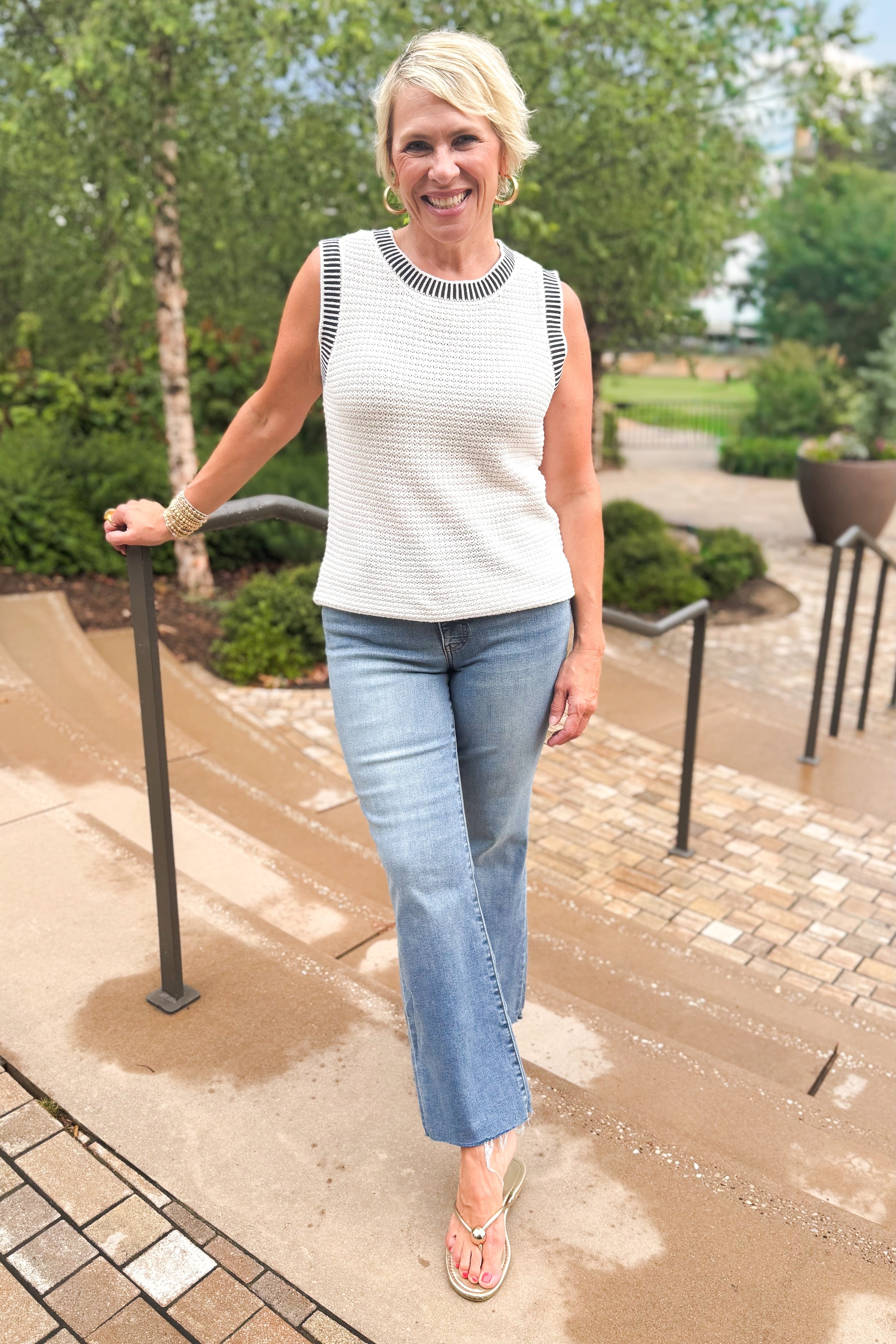 Front view of woman wearing Swagger Boutiqueβs White & Navy Textured Sweater Tank with jeans leaning against a railing outside.