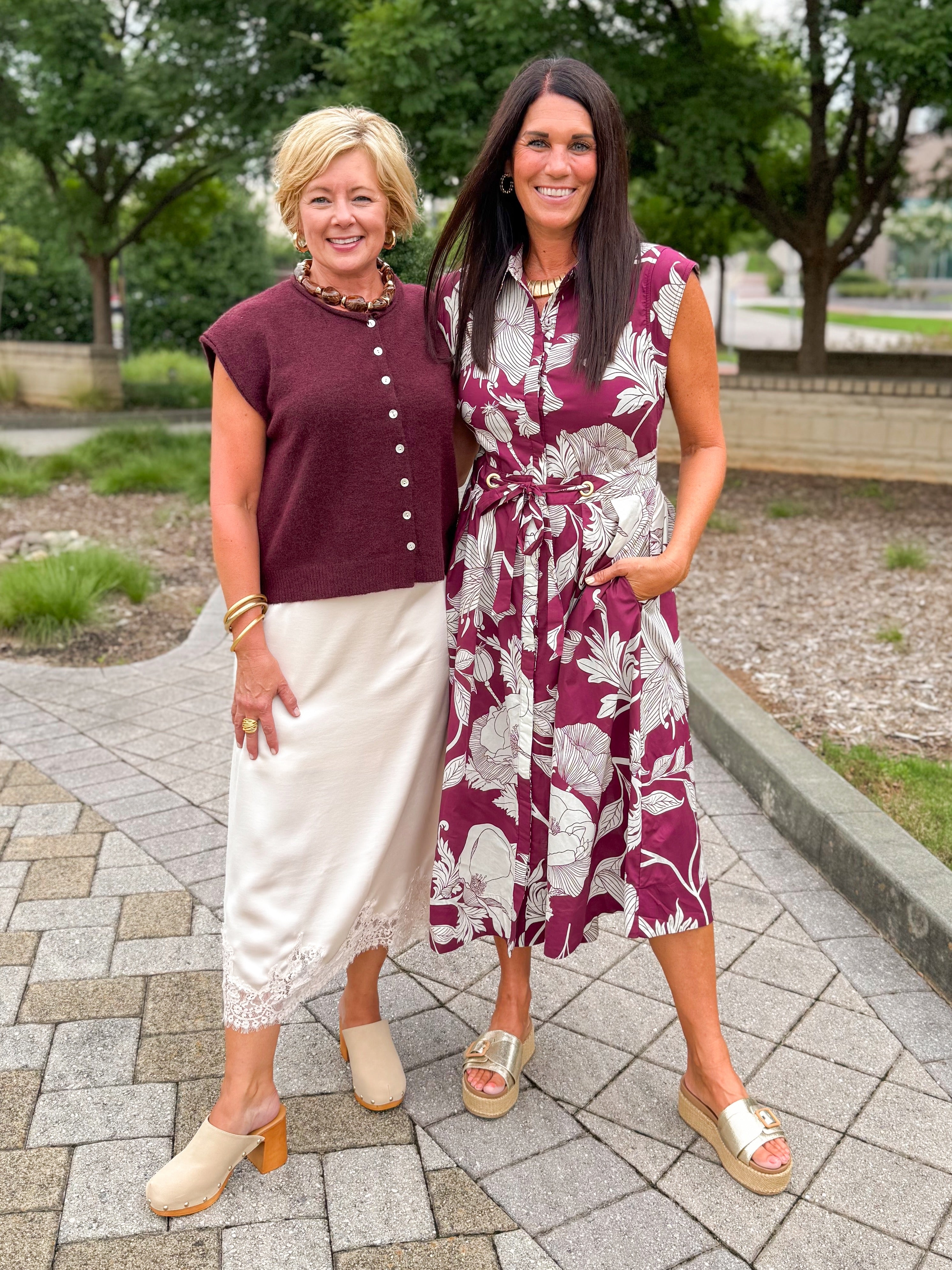 Front view of women wearing Swagger Boutiqueās Wine Sleeveless Button Up Sweater with Ecru Lace Lined Satin Skirt by Entro and floral midi dress standing outside at a park.