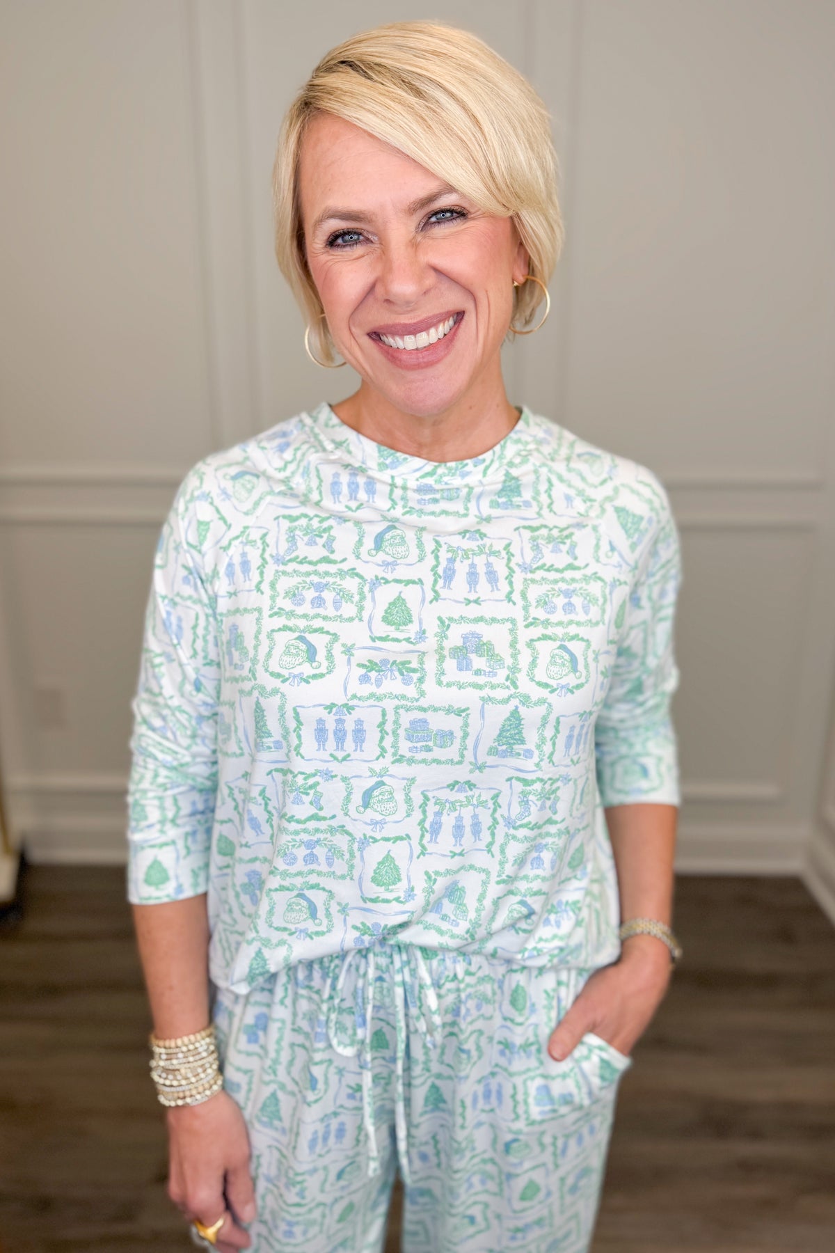 Model wearing the Yueltide Vignettes Blue Annie Pajama Set, featuring a festive light blue and green holiday print on a white background with Christmas trees, wreaths, and decorative holiday motifs. The set includes a relaxed-fit, long-sleeve crewneck top and matching straight-leg pajama pants.