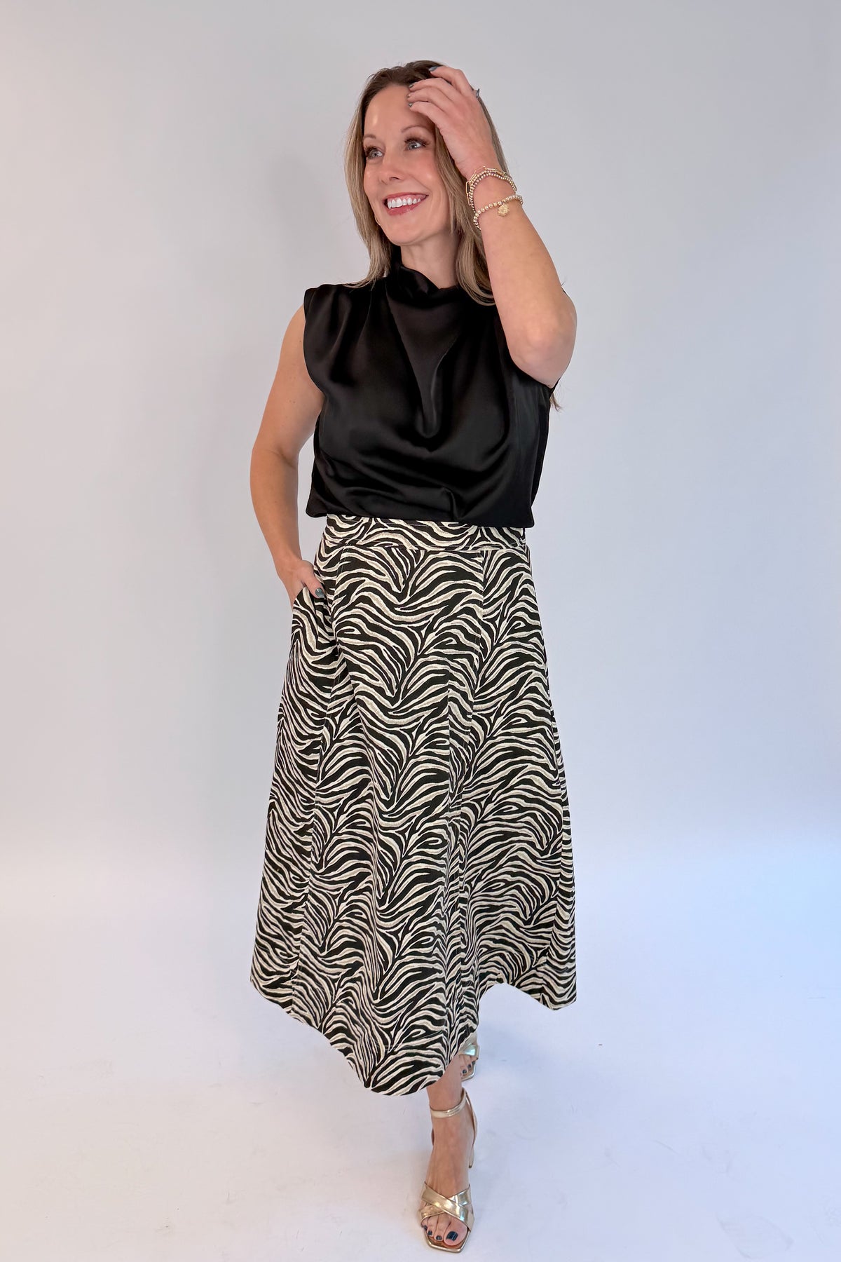 Front view of woman wearing Swagger Boutique’s Zebra Print Midi Skirt by Entro & Black Satin Cowlneck Sleeveless Top against white studio background.