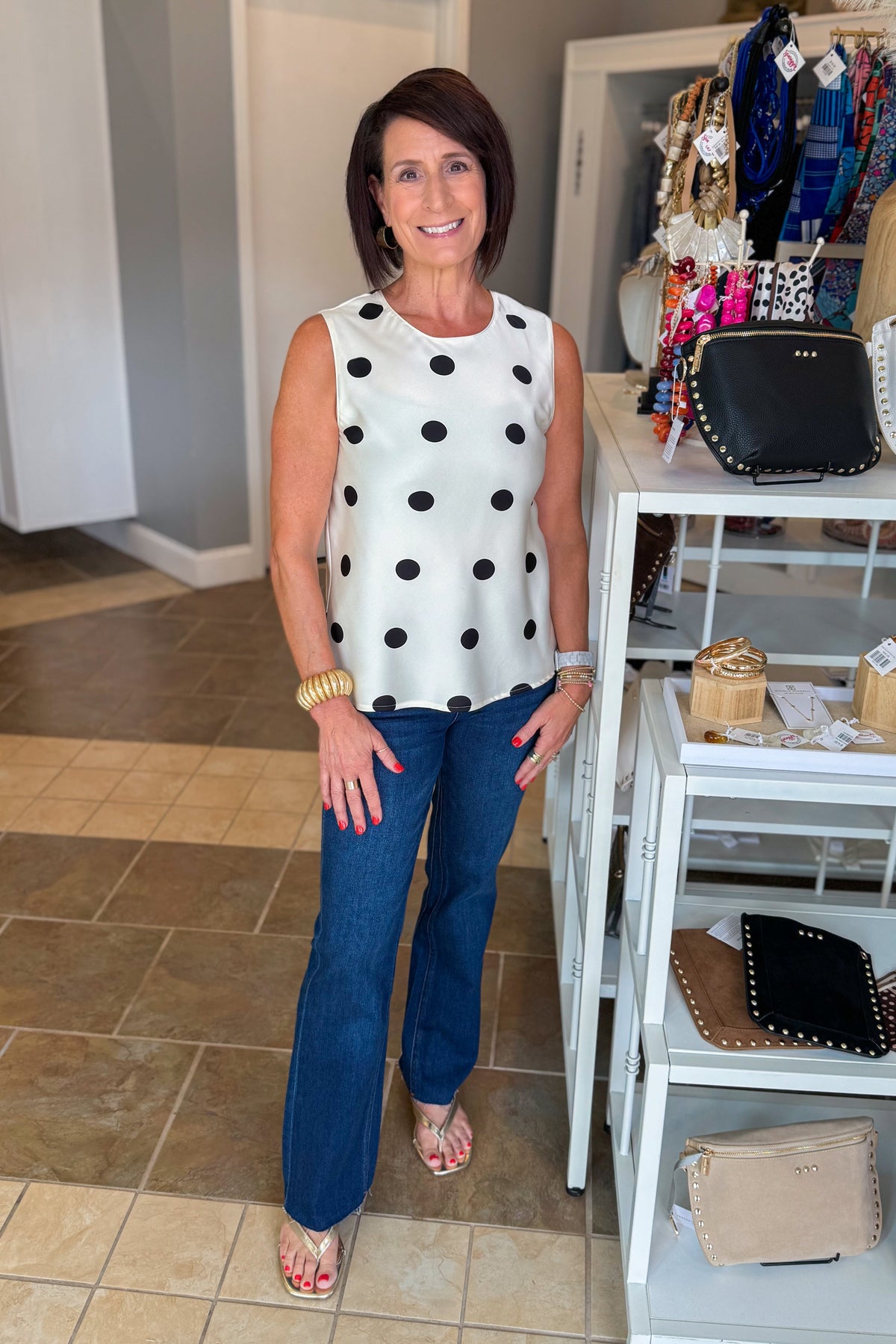 Front view of woman wearing Swagger Boutique’s Cream & Black Polka Dot Satin Tank with jeans standing at the store.