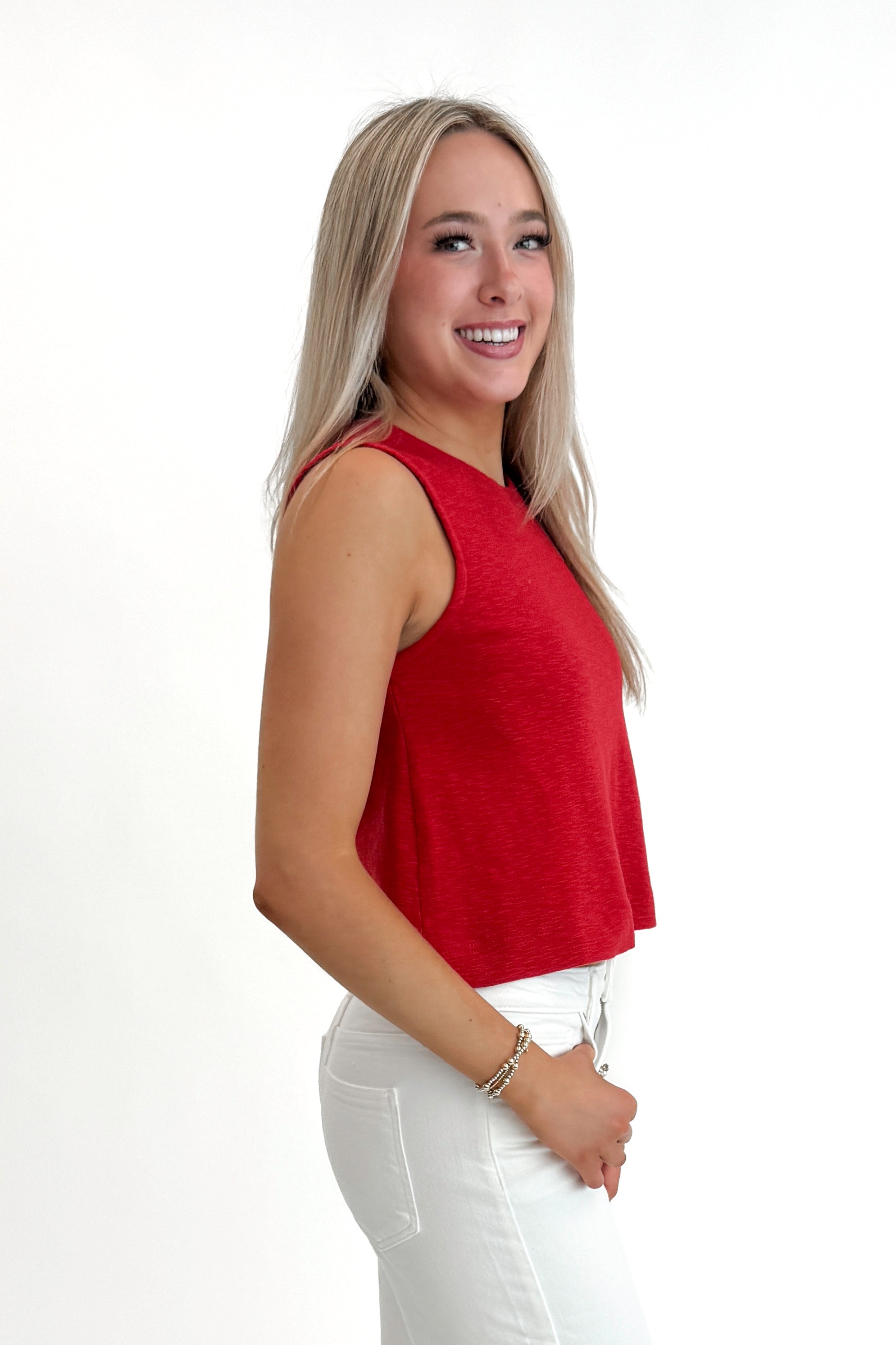 Side view of woman wearing Red Boxy Tank Top with white jeans against white studio background.