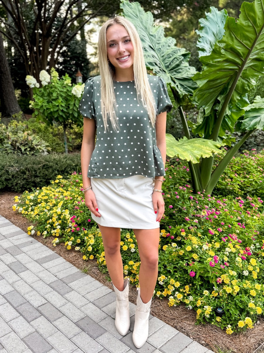 A woman stands outdoors in a garden wearing a sage green short-sleeve top with white polka dots, a white mini skirt, and white ankle boots.