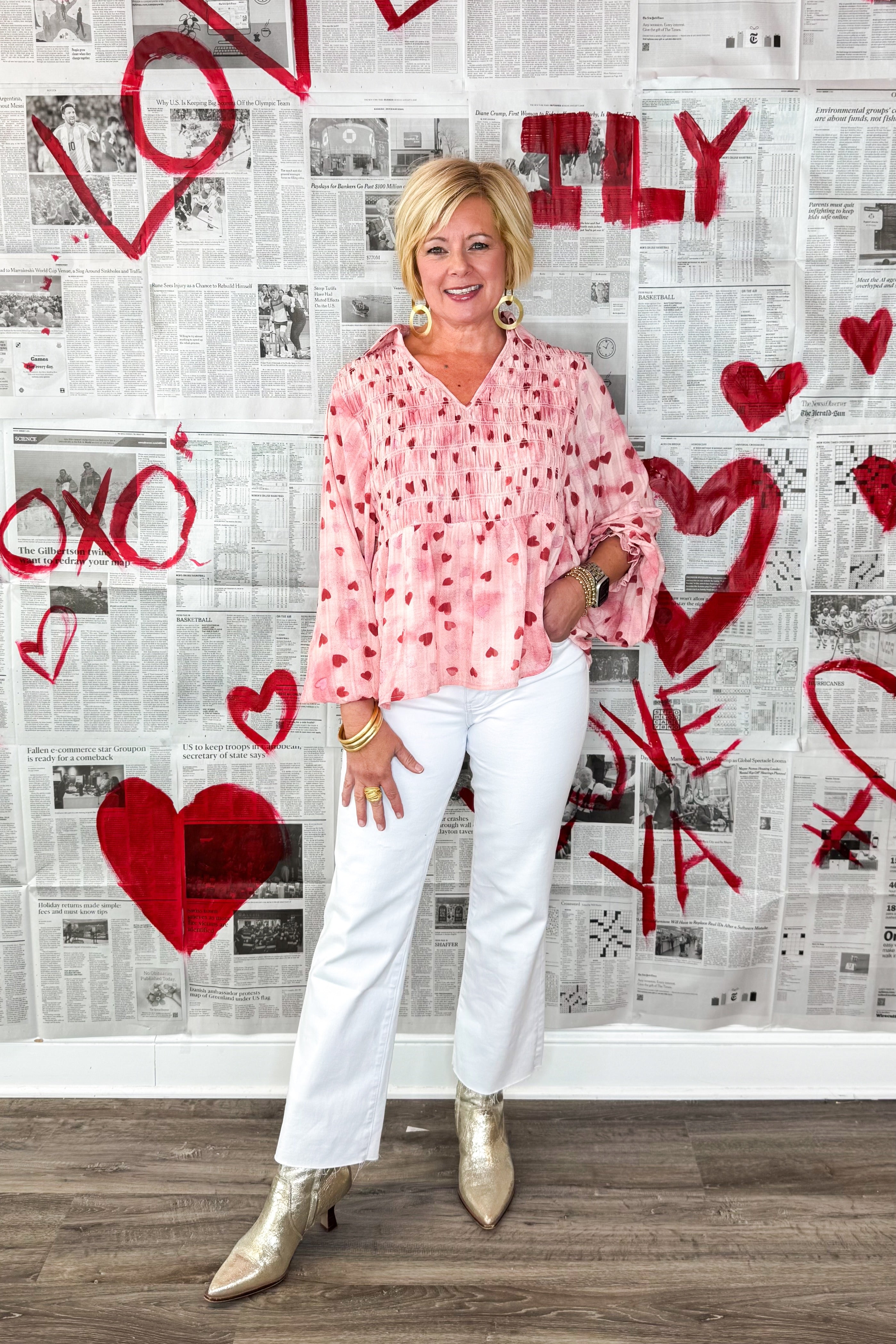 Woman in a pink blouse and white pants standing in front of a wall with red hearts and the word &