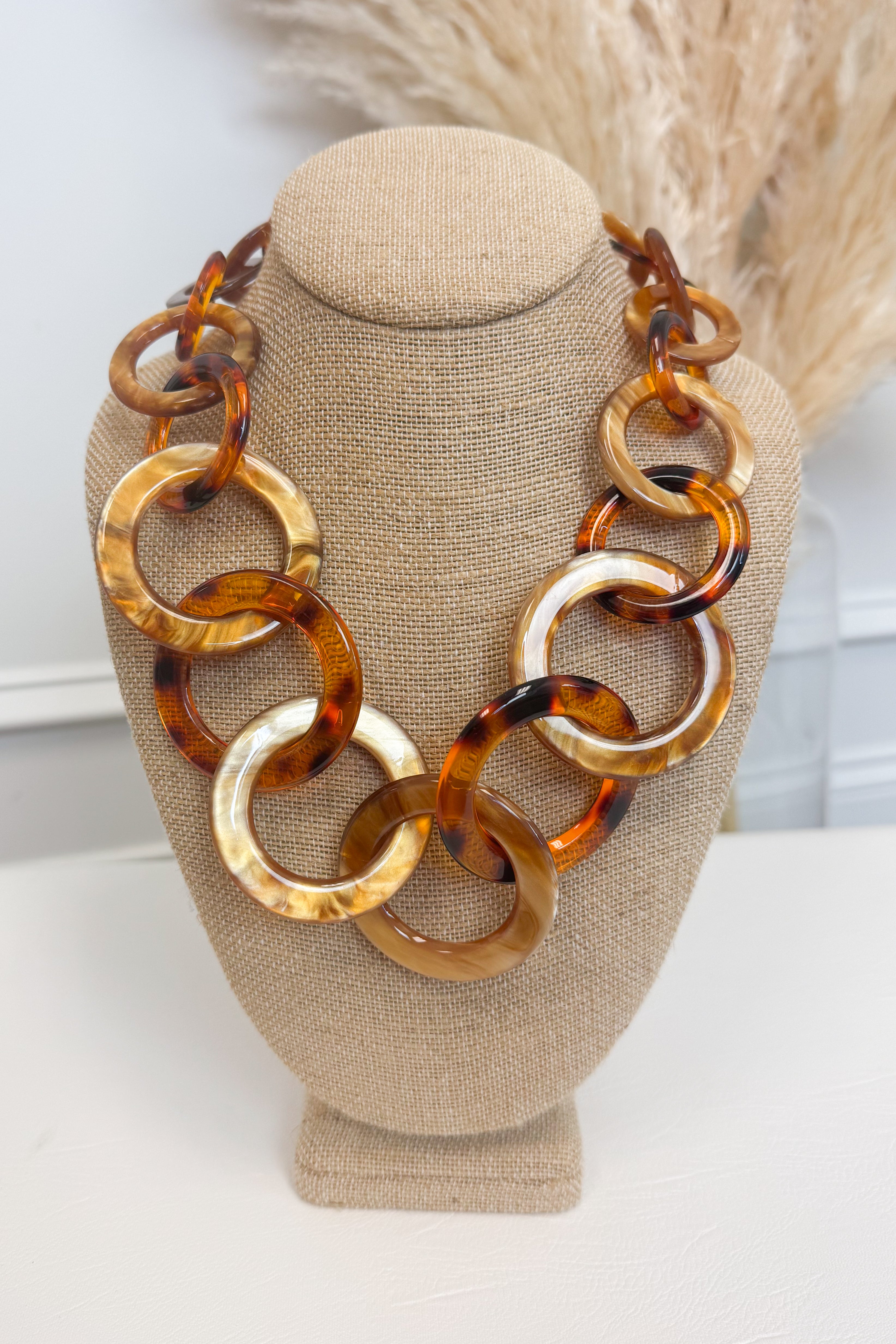 Chunky statement necklace made of large interlocking tortoiseshell and amber-colored links, displayed on a beige fabric jewelry bust against a light neutral background with dried grass decor.