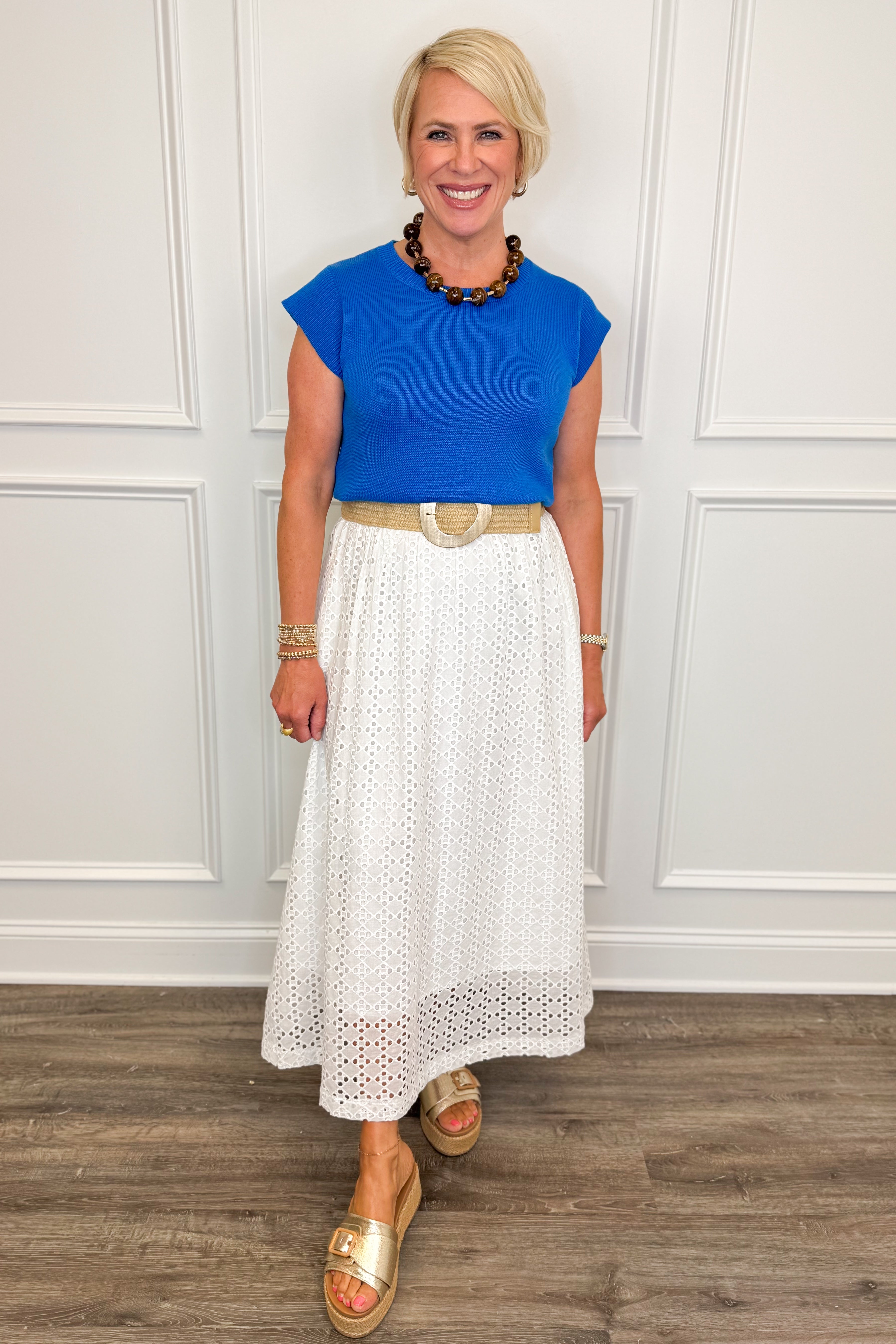 Woman wearing a blue top and white skirt with a wooden floor and white paneled wall background