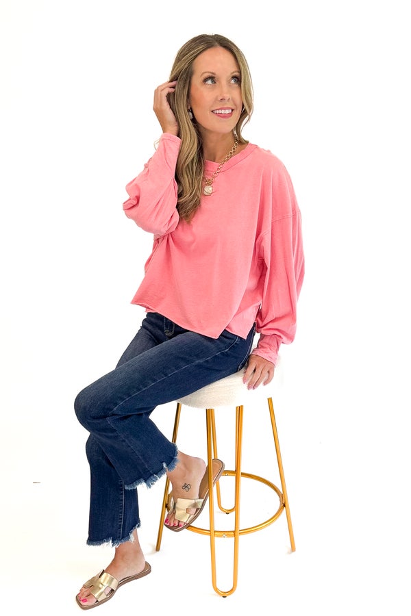Woman sitting on a stool wearing a pink top and blue jeans against a white background