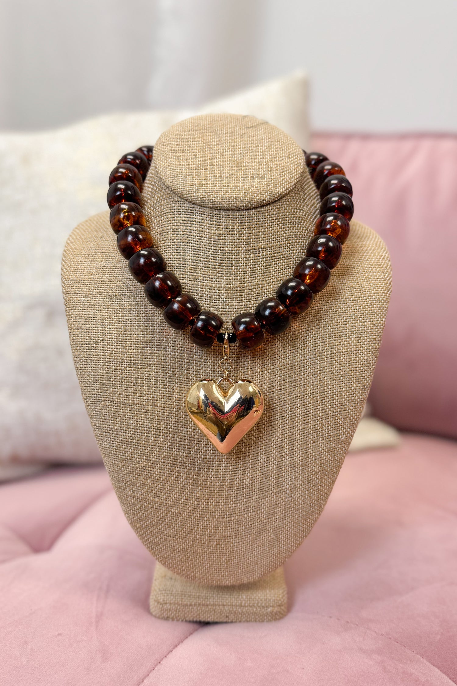 Brass beaded necklace with a heart pendant on a beige bust against a pink background