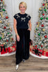 Woman in a black outfit with silver bow details standing in front of Christmas trees.