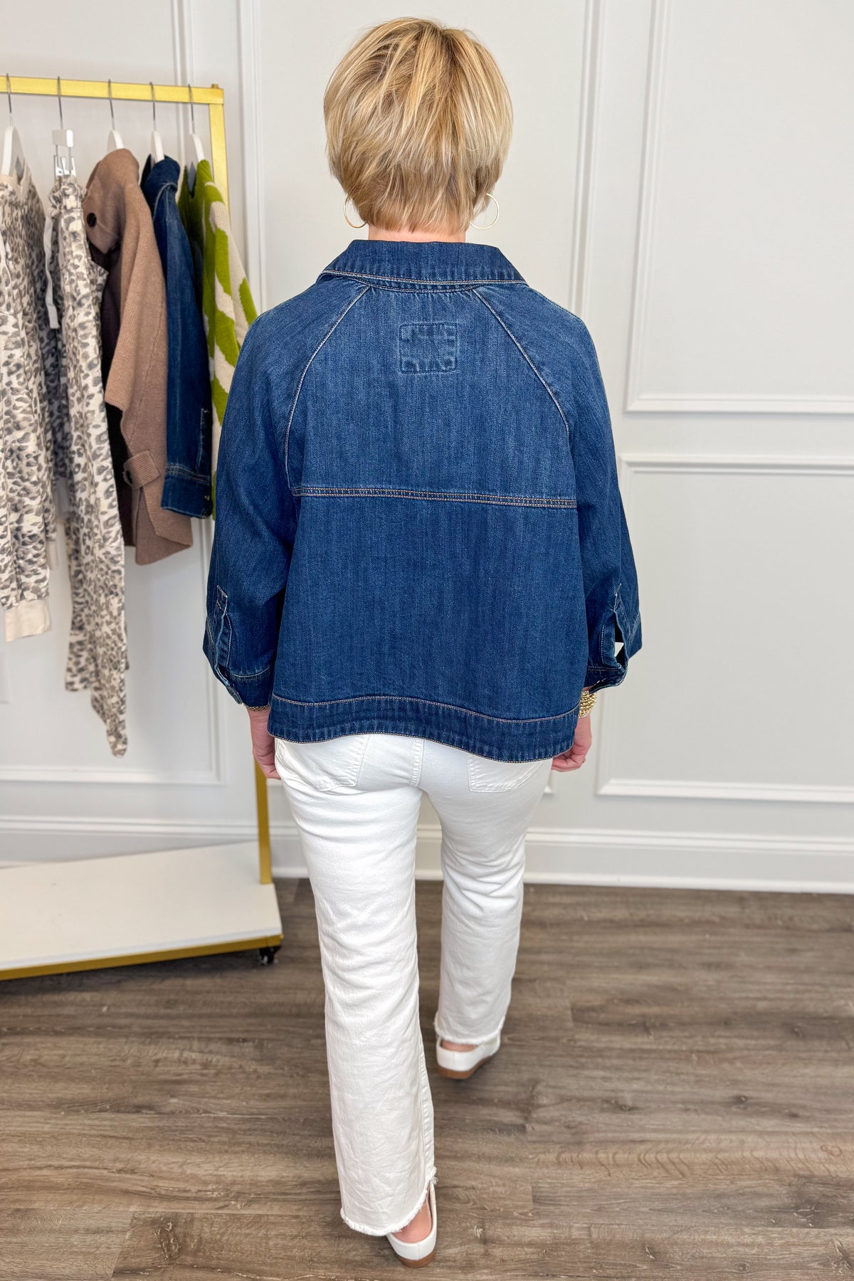 Back view of woman wearing Swagger Boutique’s Dark Denim Cape Top with jeans with clothing hanging in the background.