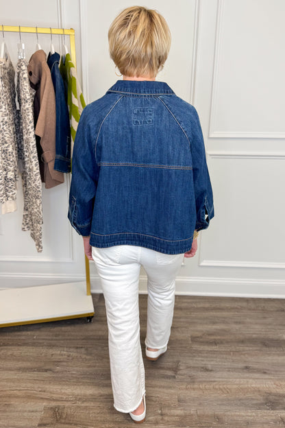 Back view of woman wearing Swagger Boutique’s Dark Denim Cape Top with jeans with clothing hanging in the background.
