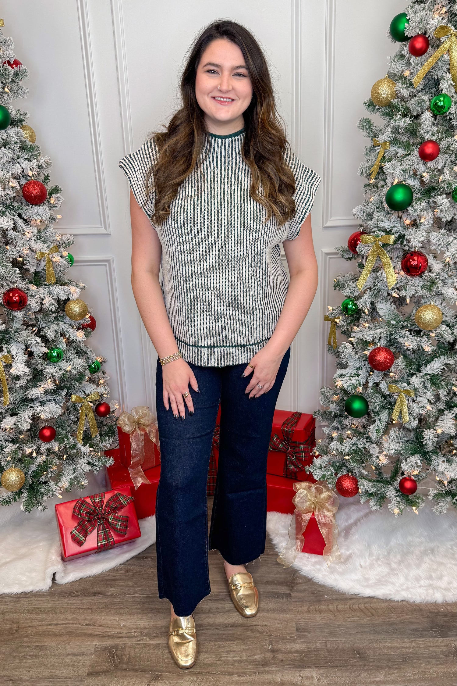 Front view of woman wearing green two toned sweater with jeans and Christmas trees in the background.