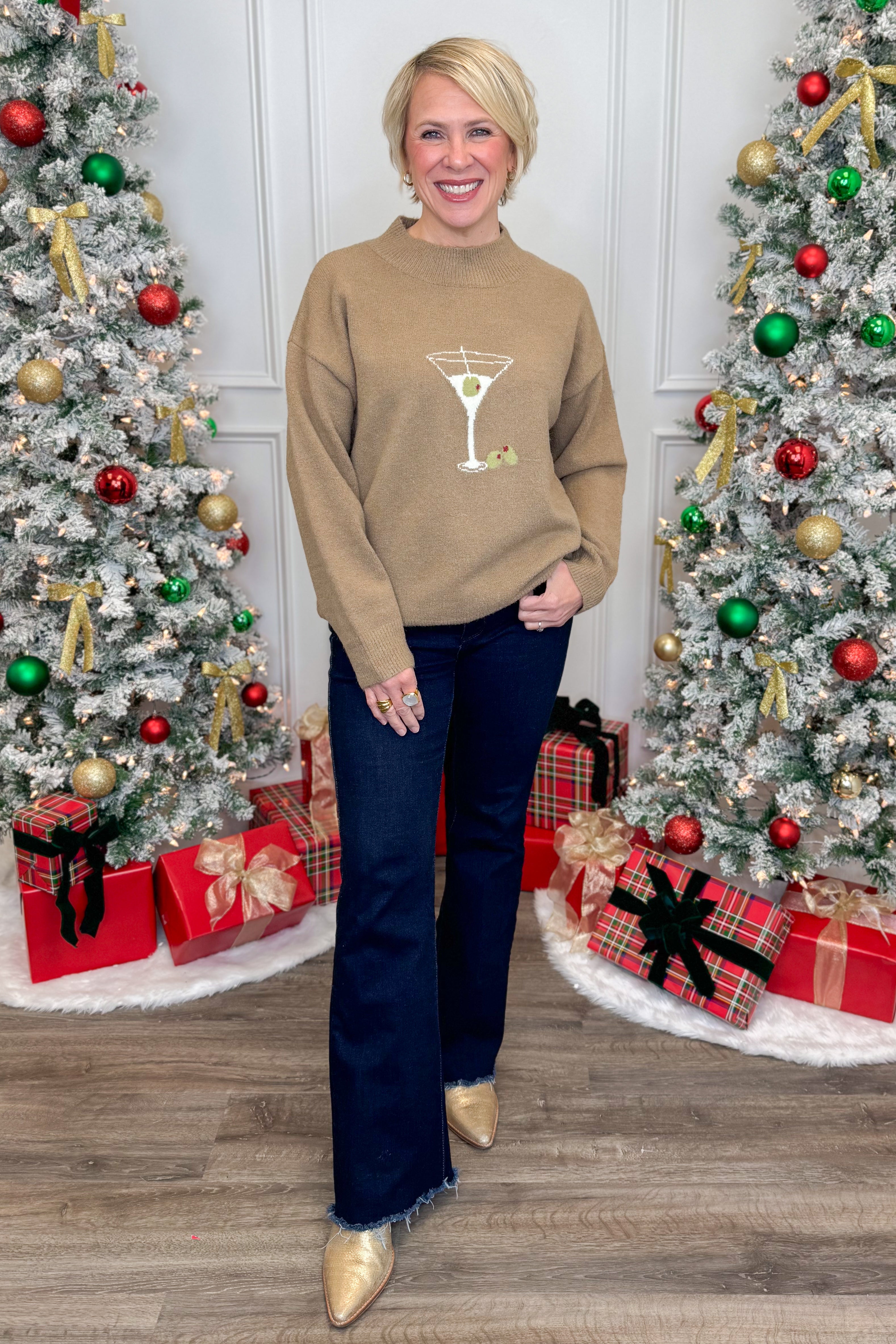 Woman wearing a brown sweater with a martini glass design in front of Christmas trees and presents.