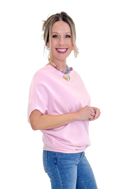 Side view of woman wearing Light Pink Dolman Sleeve Top with jeans against white studio background.