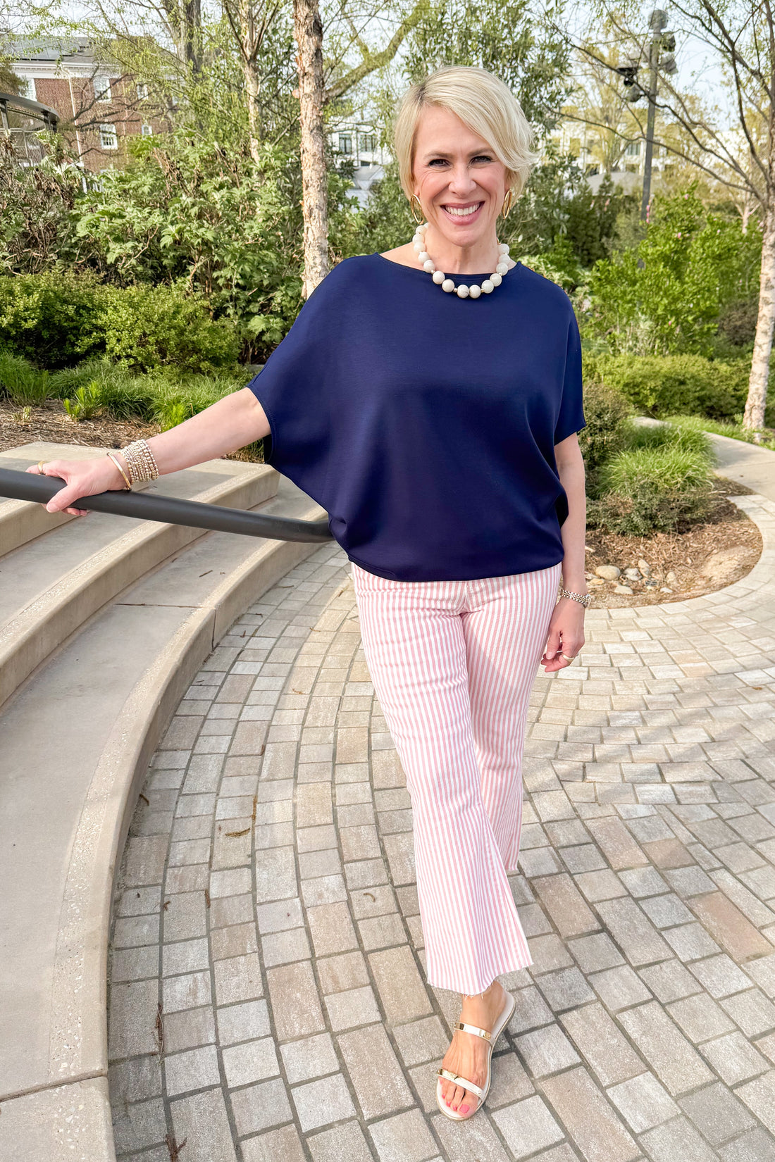 Front view of woman wearing Navy Dolman Sleeve Top with striped pink jeans outside at a park.