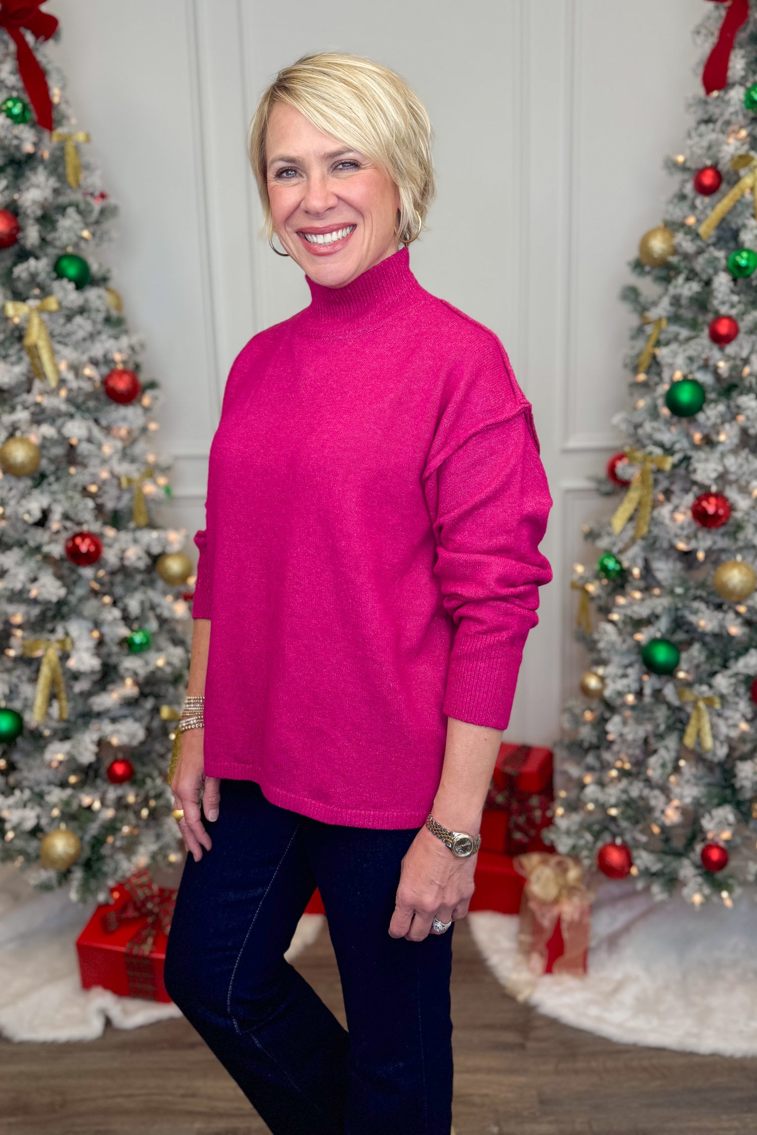 Woman in a pink sweater standing in front of Christmas trees with decorations.
