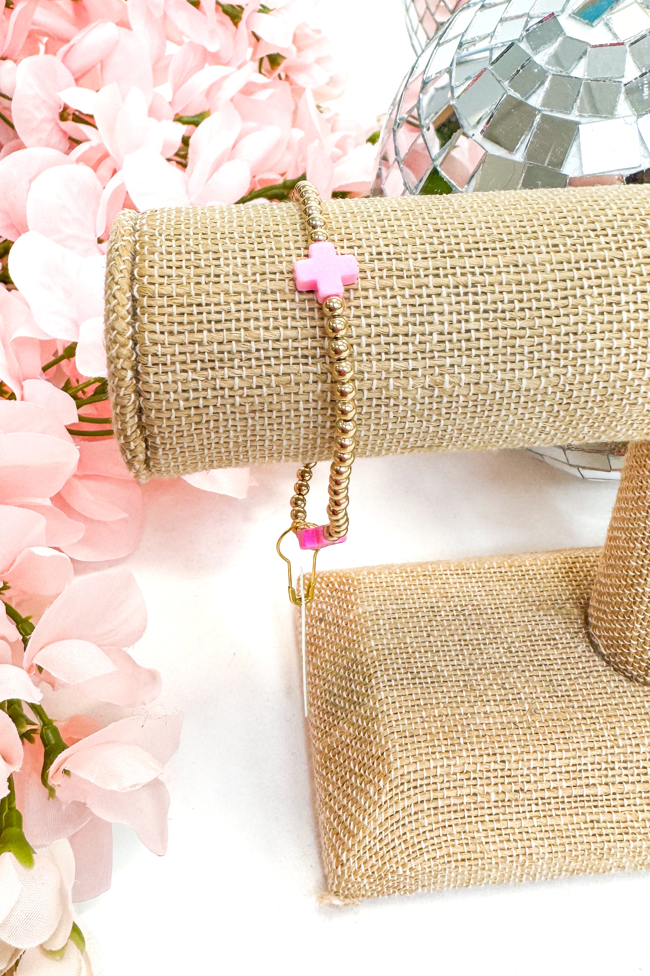 gold enewton bracelet with pink cross detailing  on burlap display with floral background