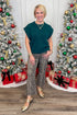 Woman in a green top and patterned pants standing between two decorated Christmas trees with presents.
