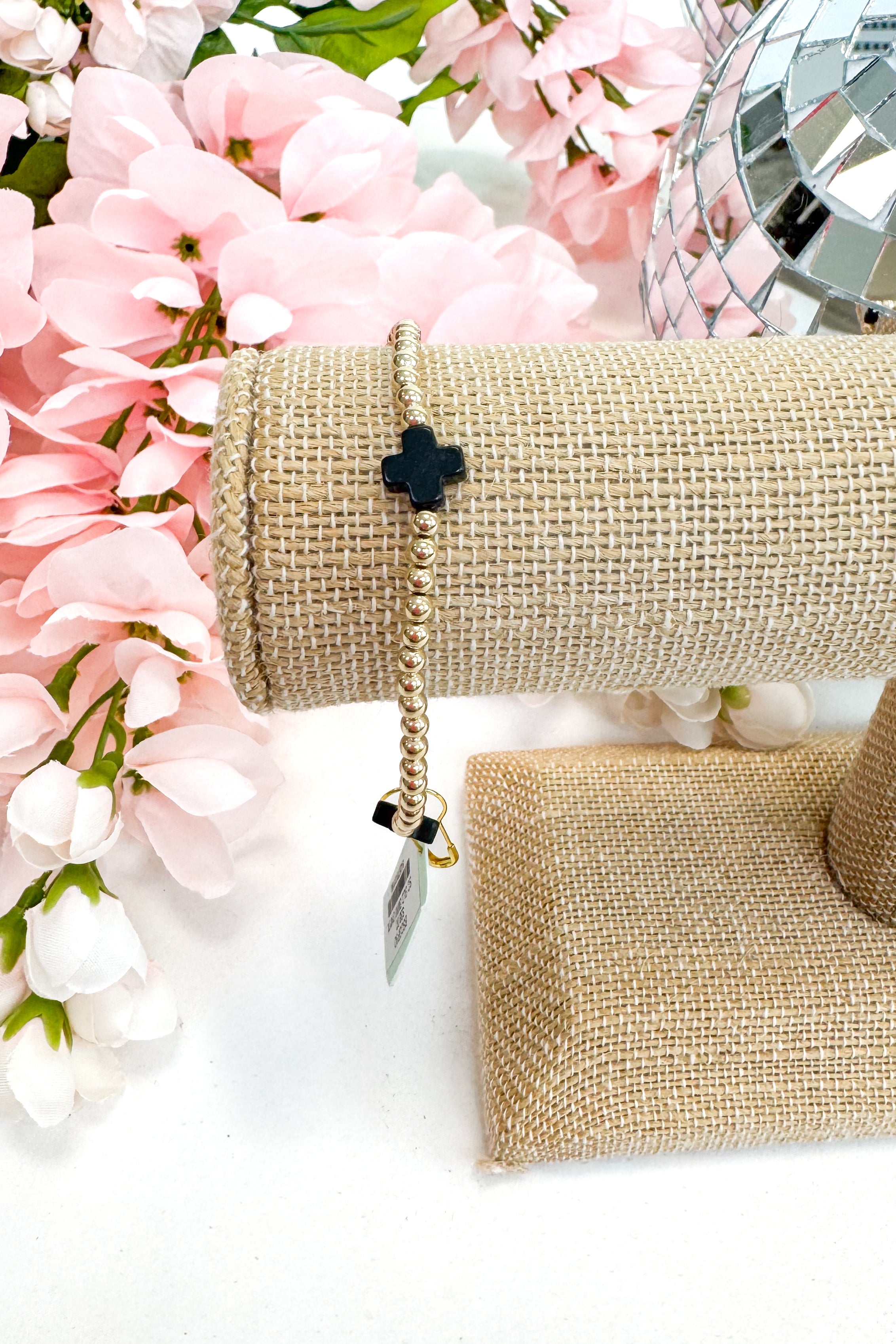 gold enewton bracelet with black cross detailing on burlap display with floral background