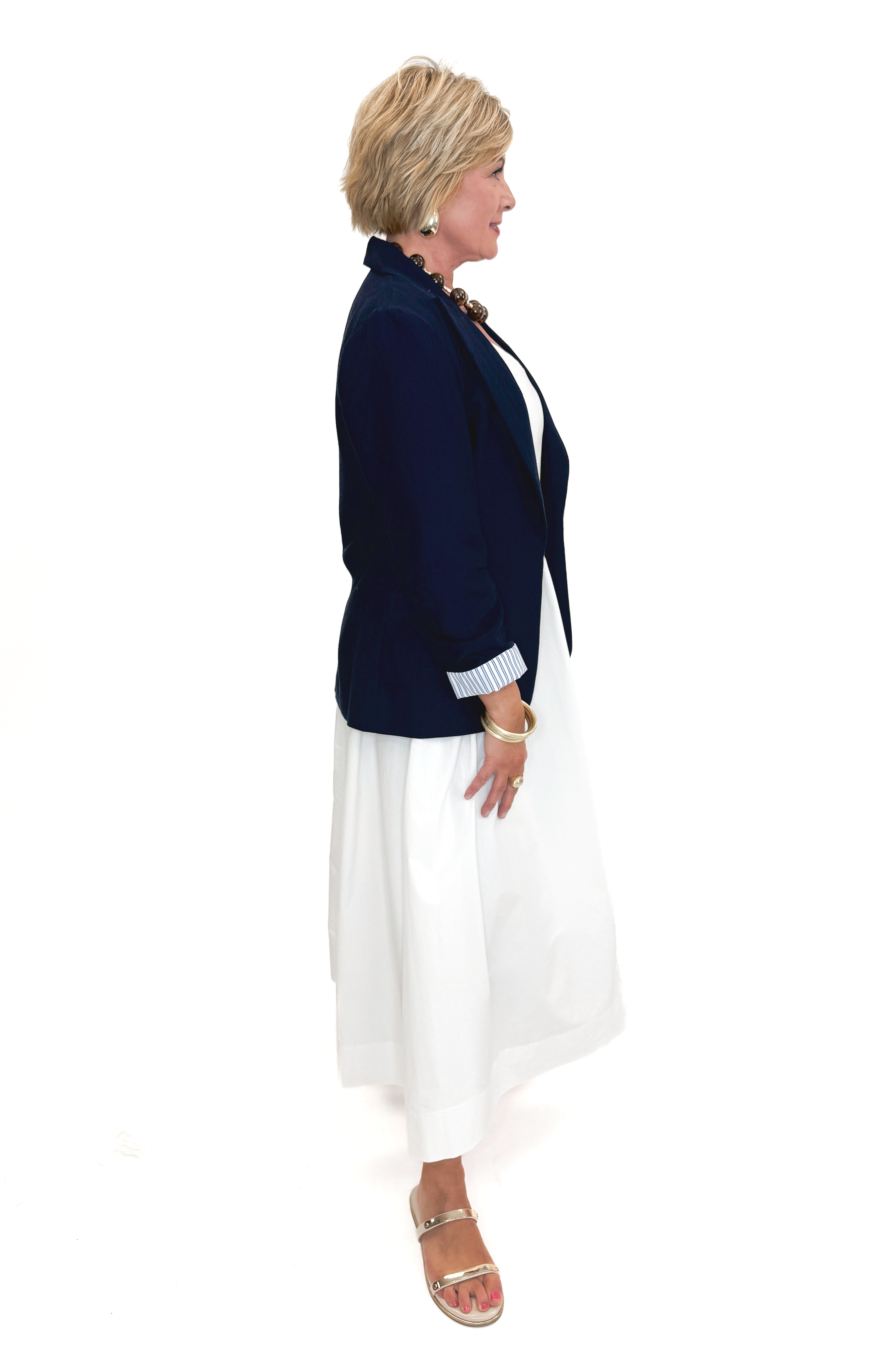 Side view of woman wearing Navy Cinched Sleeve Blazer by Skies Are Blue with white dress against white studio background.
