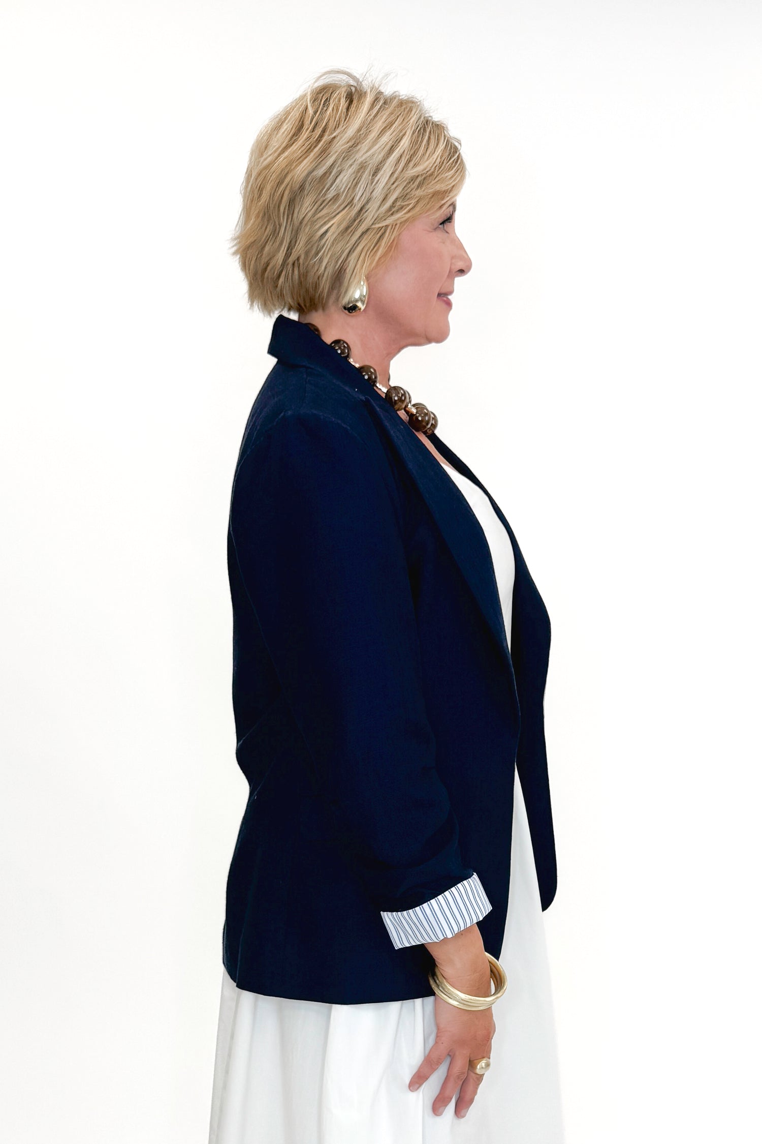 Side view of woman wearing Navy Cinched Sleeve Blazer by Skies Are Blue with white dress against white studio background.