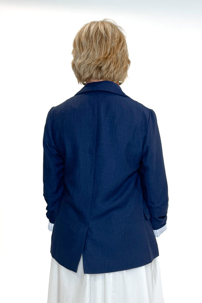 Back view of woman wearing Navy Cinched Sleeve Blazer by Skies Are Blue with white dress against white studio background.