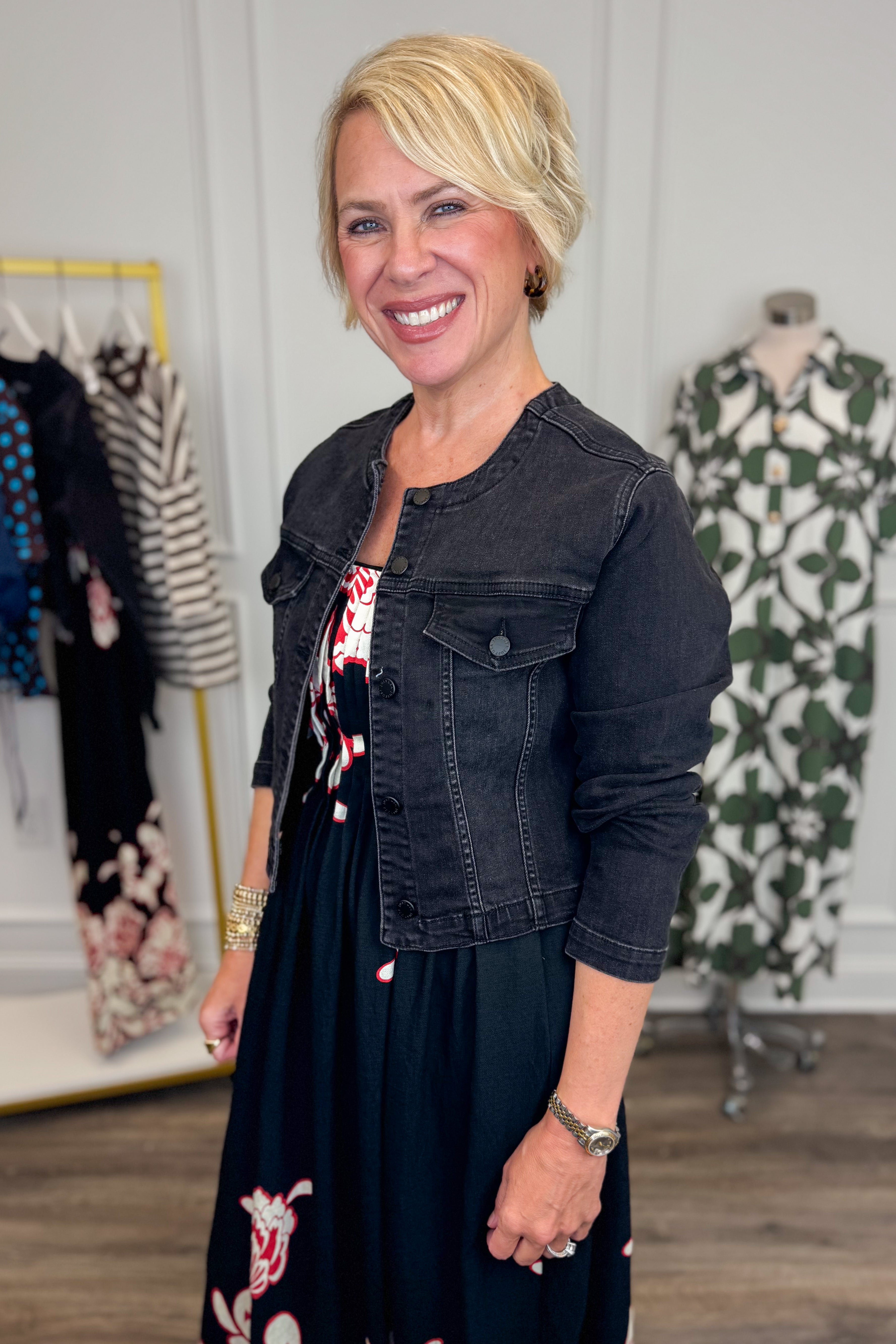 Side view of woman wearing Swagger Boutique’s washed black denim jacked with dress with clothing hanging in the background.