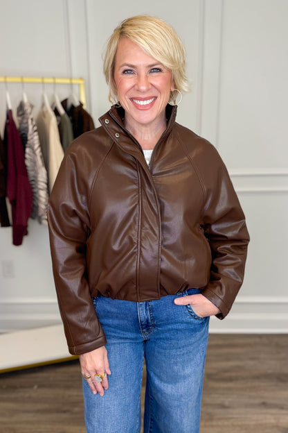 A smiling blonde woman wearing a brown faux-leather zip-front jacket with a high collar and snap details, paired with blue jeans, standing in a boutique setting.