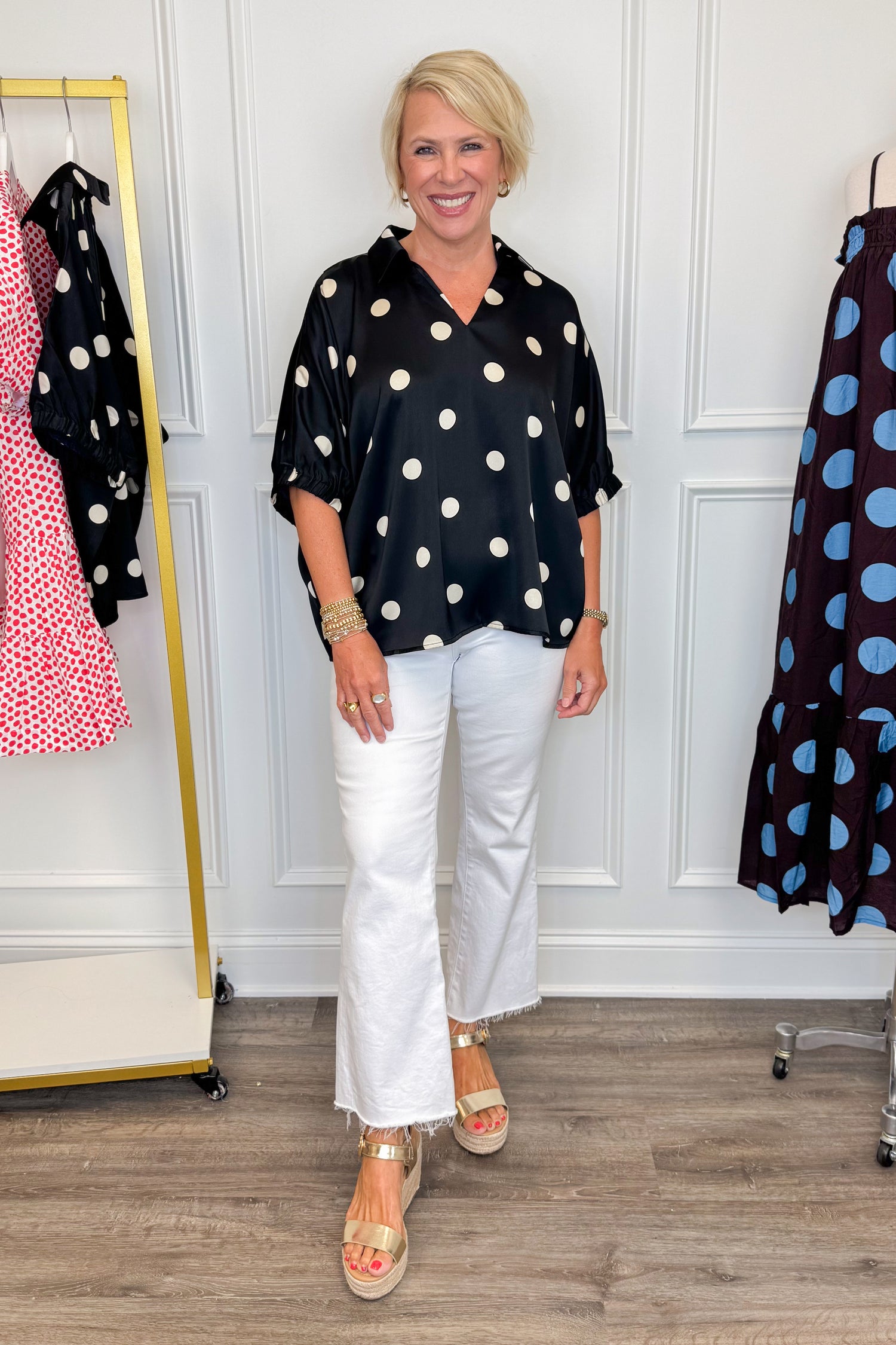 Front view of woman wearing Swagger Boutique’s Black &amp; Cream Polka Dot Satin Top with jeans with clothing hanging in the background.