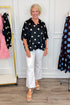 Front view of woman wearing Swagger Boutique’s Black & Cream Polka Dot Satin Top with jeans with clothing hanging in the background.