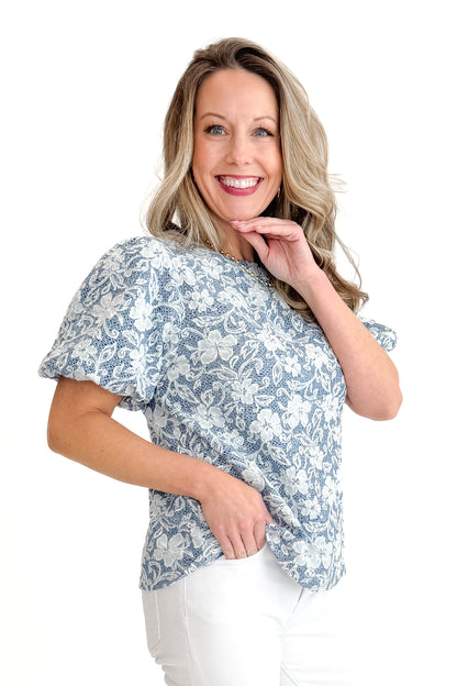 Side view of woman wearing Light Blue Floral Textured Top with jeans against white studio background.