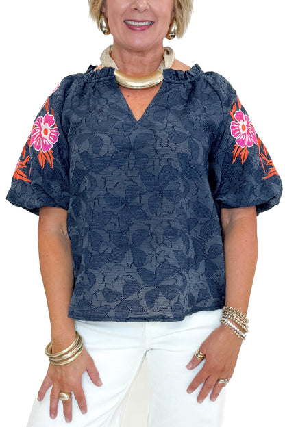 Front detail view of woman wearing Navy With Floral Embroidered Sleeve Top with jeans against white studio background.