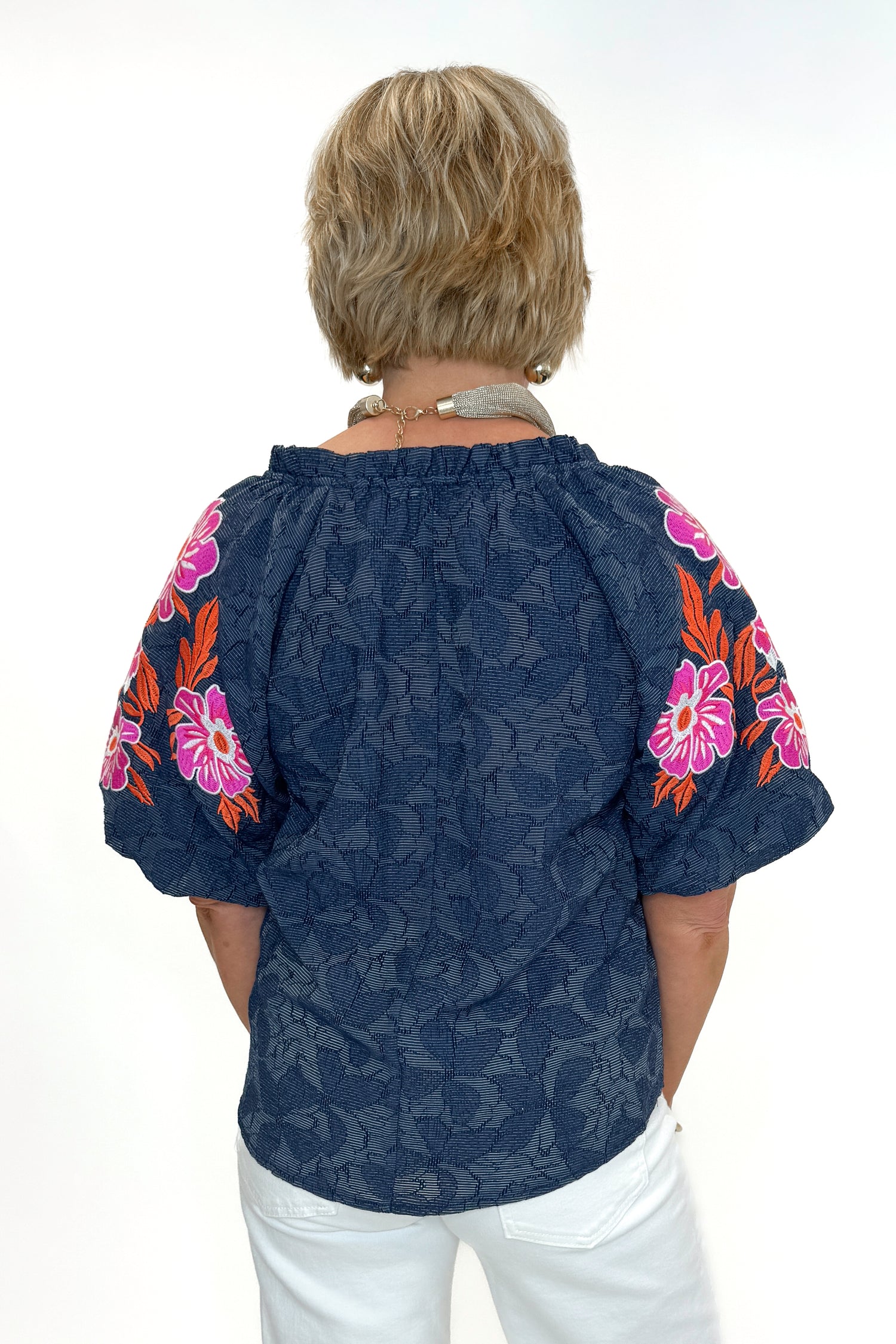 Back view of woman wearing Navy With Floral Embroidered Sleeve Top with jeans against white studio background.