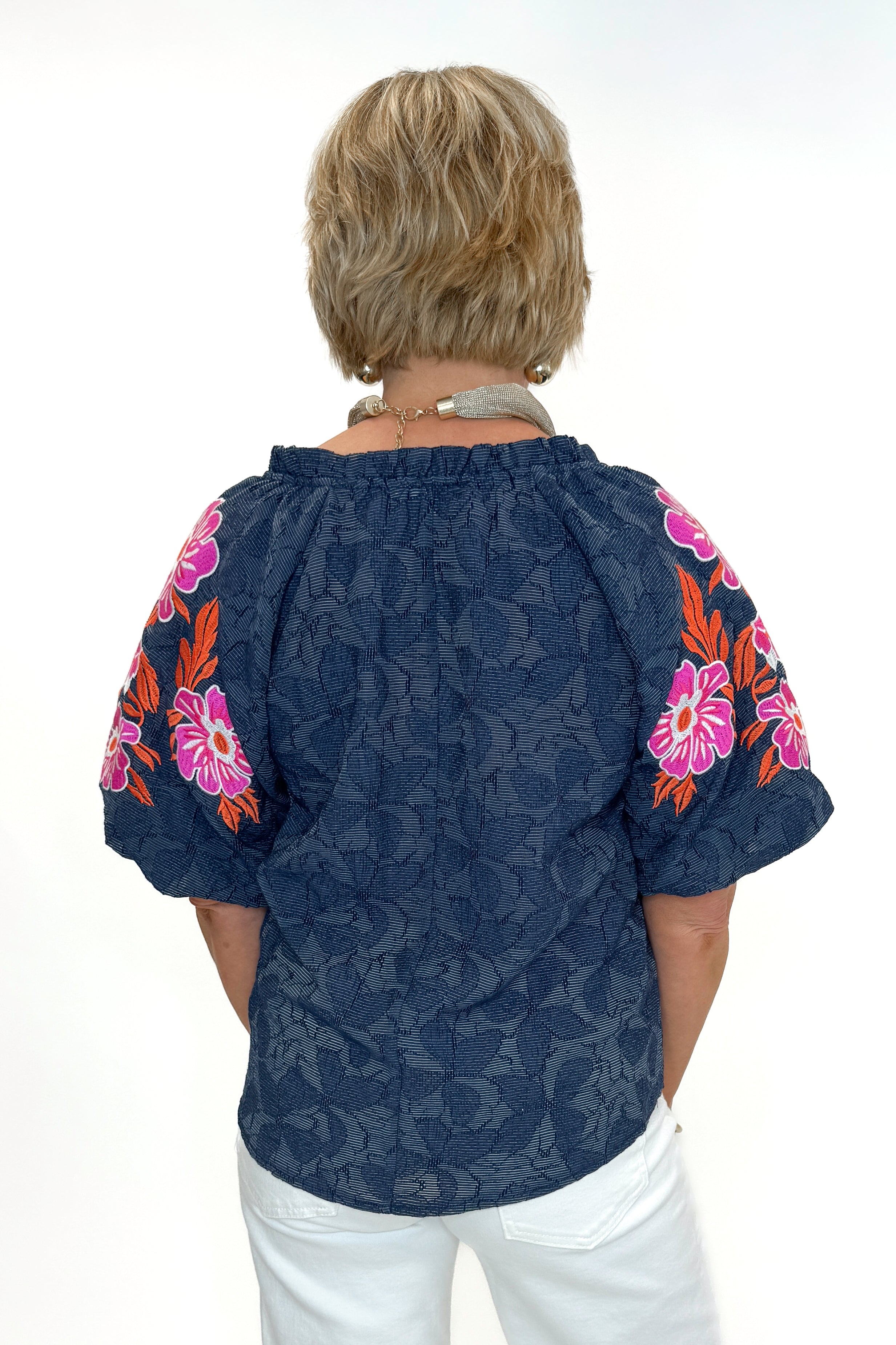 Back view of woman wearing Navy With Floral Embroidered Sleeve Top with jeans against white studio background.